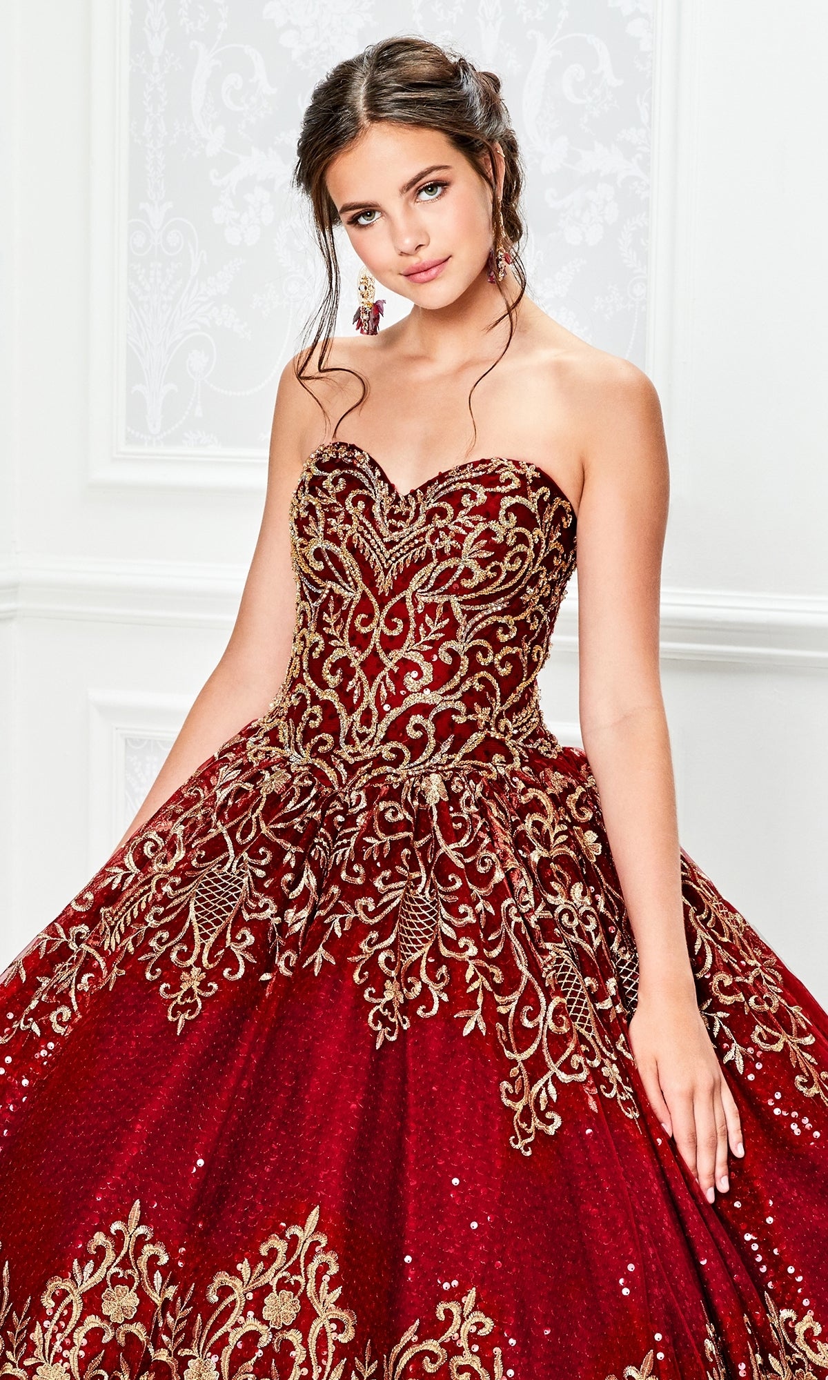 A young woman wears the Princesa Red and Gold Quinceañera Dress PR11945, a strapless ball gown with gold embroidery and sequin details, standing indoors against an ornate white background.