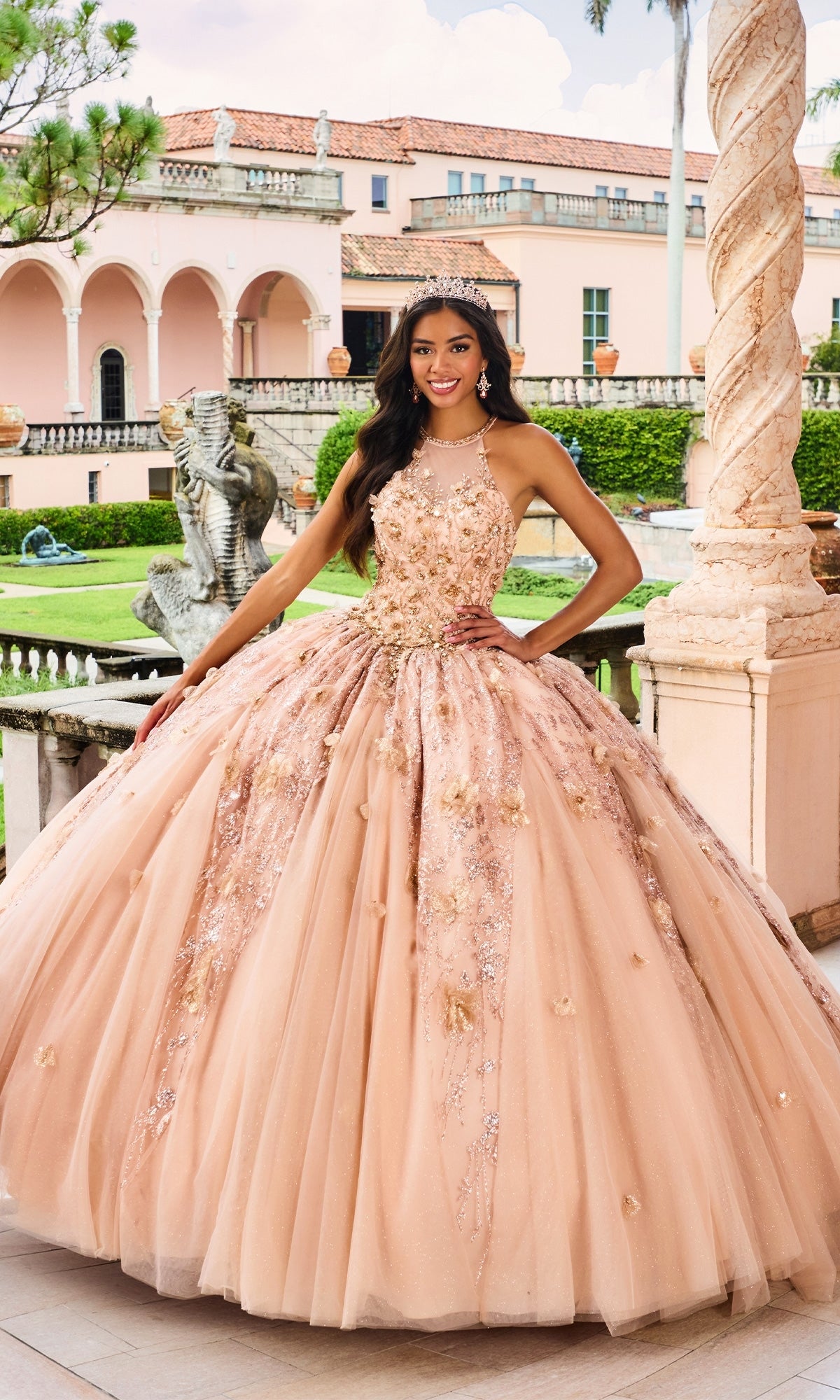 A young woman poses outdoors in the High-Neck Princesa Quinceañera Dress PR12262, her sparkling beaded peach ballgown and tiara glowing against an elegant pink historic building with arches and statues.