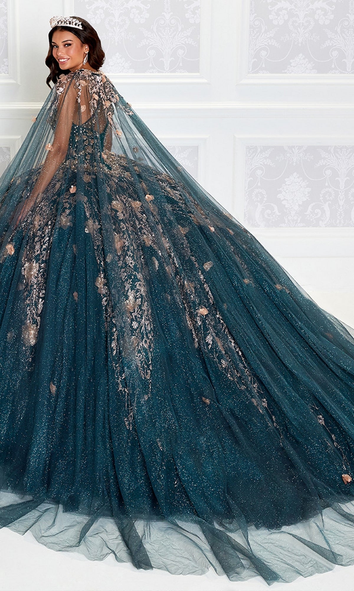 A woman stands in an elegant, white-paneled room wearing the High-Neck Princesa Quinceañera Dress PR12262, a dark teal beaded gown with glitter, floral embellishments, cape, and tiara—radiating regal style.