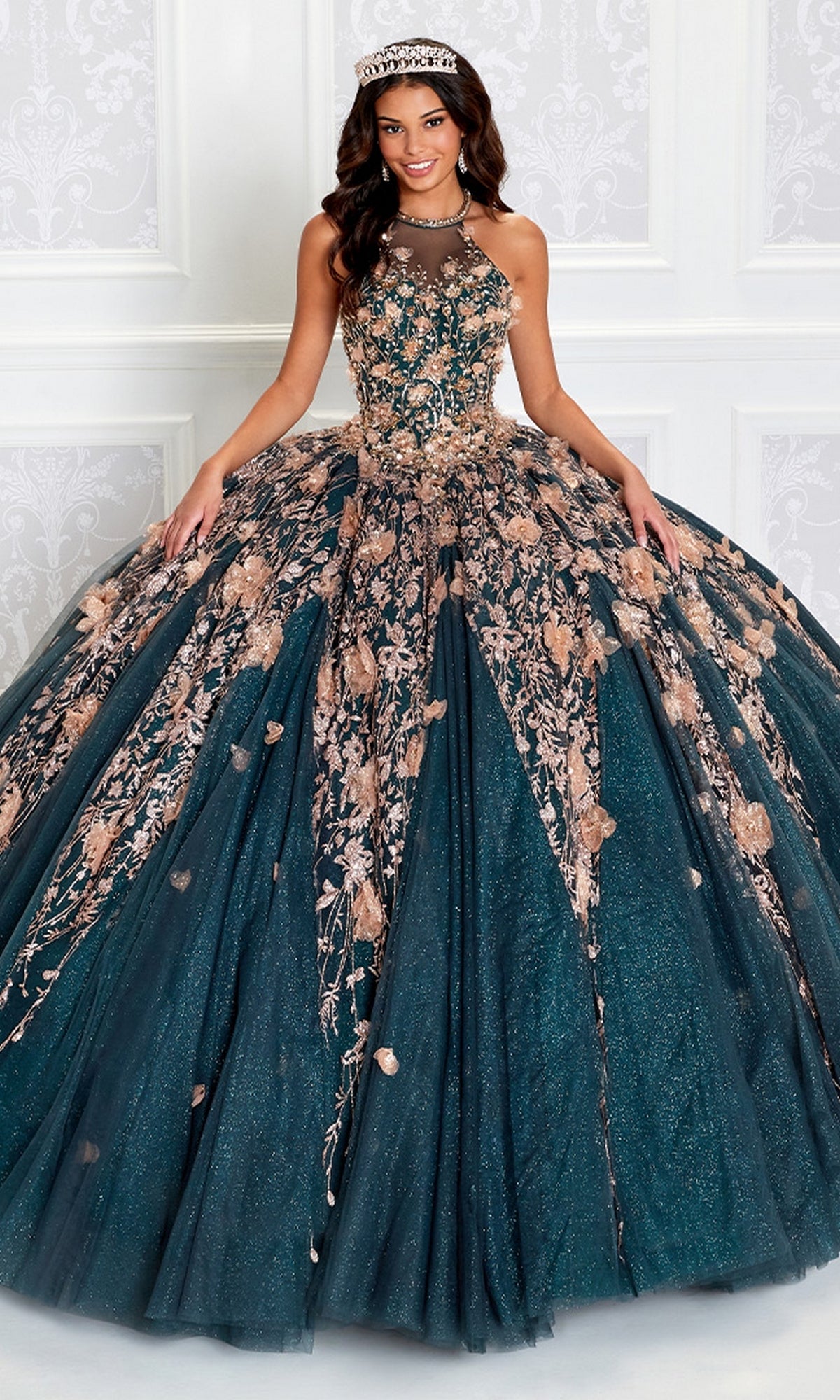 A young woman stands before a white ornate wall, wearing the High-Neck Princesa Quinceañera Dress PR12262—a dark teal ball gown with gold floral embroidery, jeweled tiara, and a full skirt.