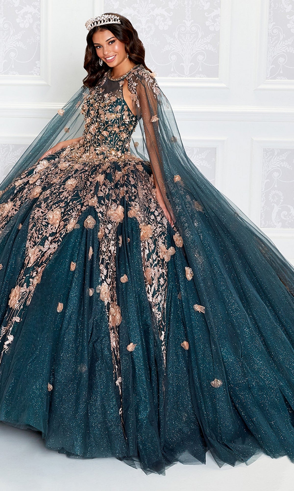 A woman smiles in the High-Neck Princesa Quinceañera Dress PR12262, featuring sparkling teal fabric, gold floral embroidery, a sheer cape, and a tiara—an enchanting look inspired by Ariana Vara.