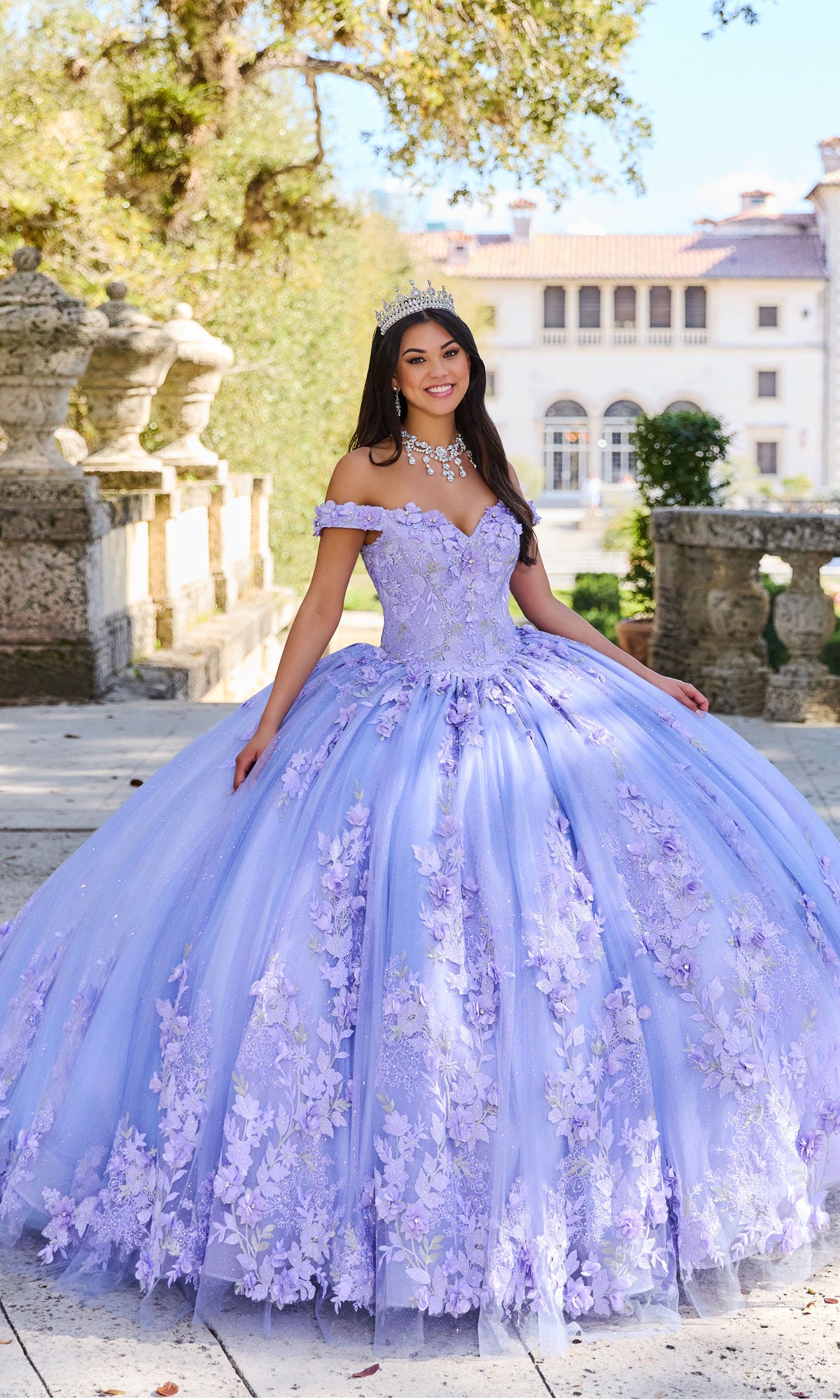 A young woman in the Princesa Quinceañera Dress PR12263 with 3-D flowers and a tiara stands outdoors in front of a mansion on a sunny day.
