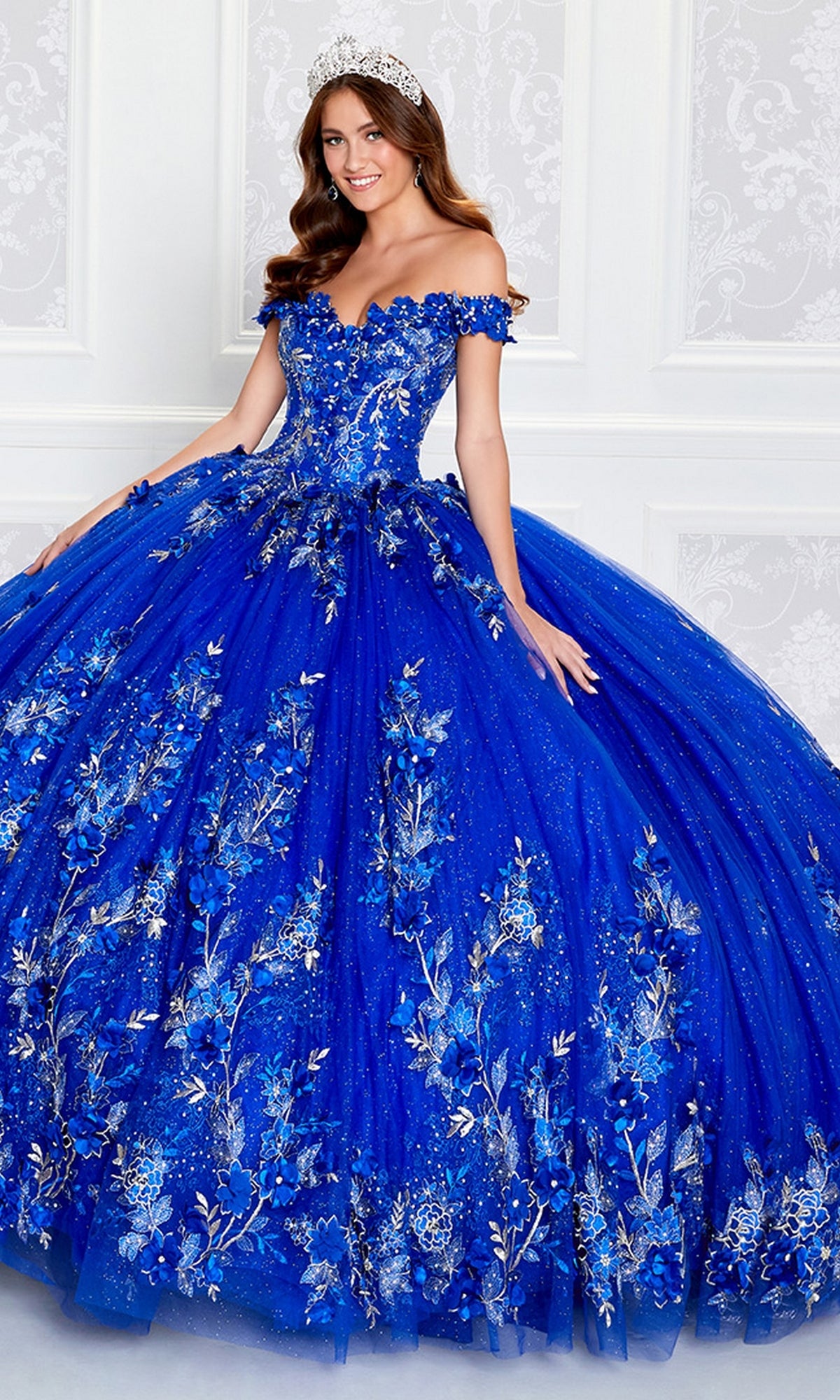 A woman in the Princesa Quinceañera Dress PR12263 with 3-D Flowers in royal blue, featuring silver floral embellishments and an off-the-shoulder design, poses in front of a white decorative background wearing a tiara.