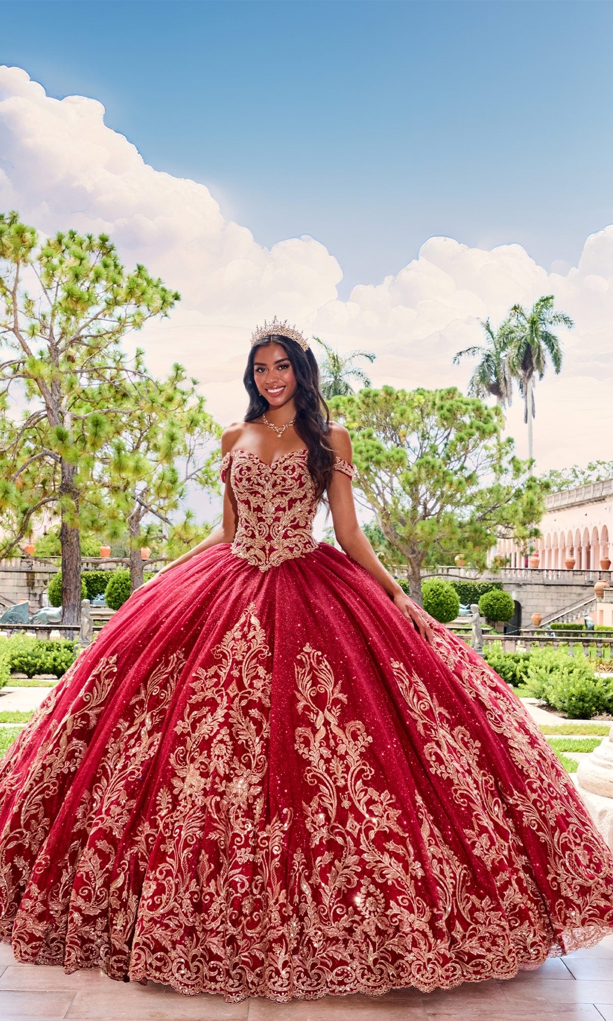 A young woman wearing a crown and the PR12264 Princesa Quince Dress with a glitter tulle skirt stands outdoors in a formal garden, with trees and classical architecture in the background.