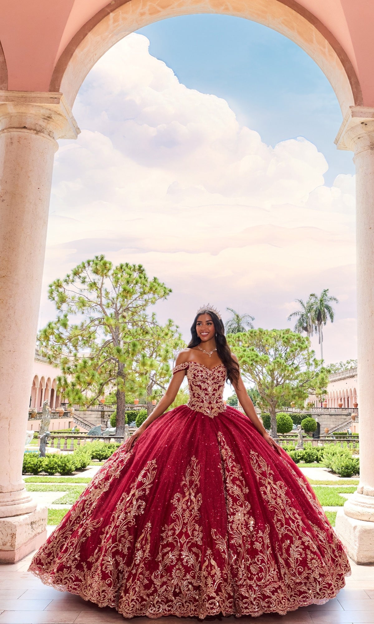 A young woman smiles at the camera in an outdoor garden, wearing the PR12264 Princesa Quince Dress with gold embroidery and a glitter tulle skirt, paired with a tiara, standing between stone columns.