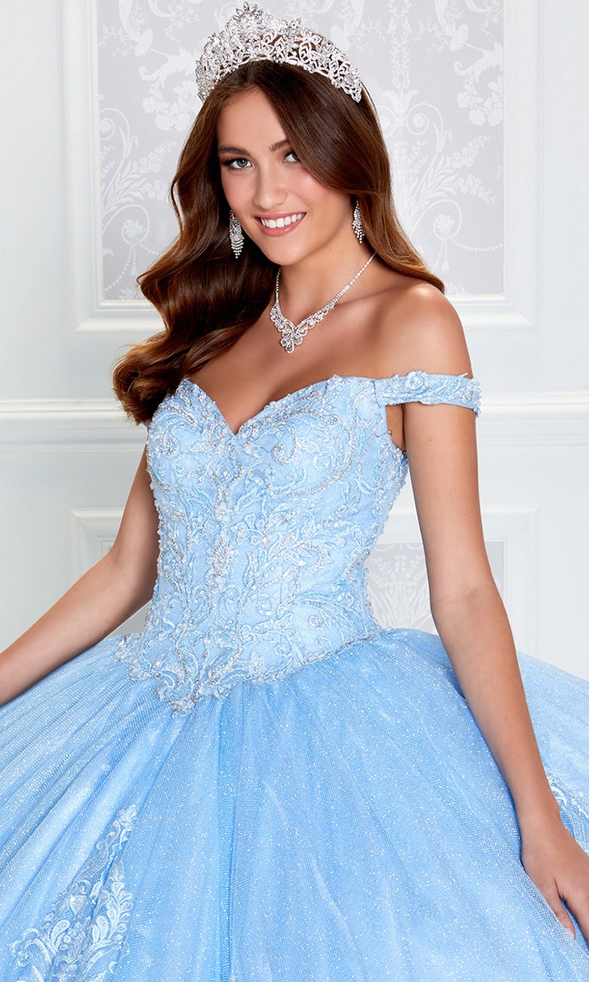 Young woman wearing the PR12264 Princesa Quince Dress with Glitter Tulle, featuring intricate embroidery, a silver tiara, and matching necklace, standing indoors against a white detailed backdrop.
