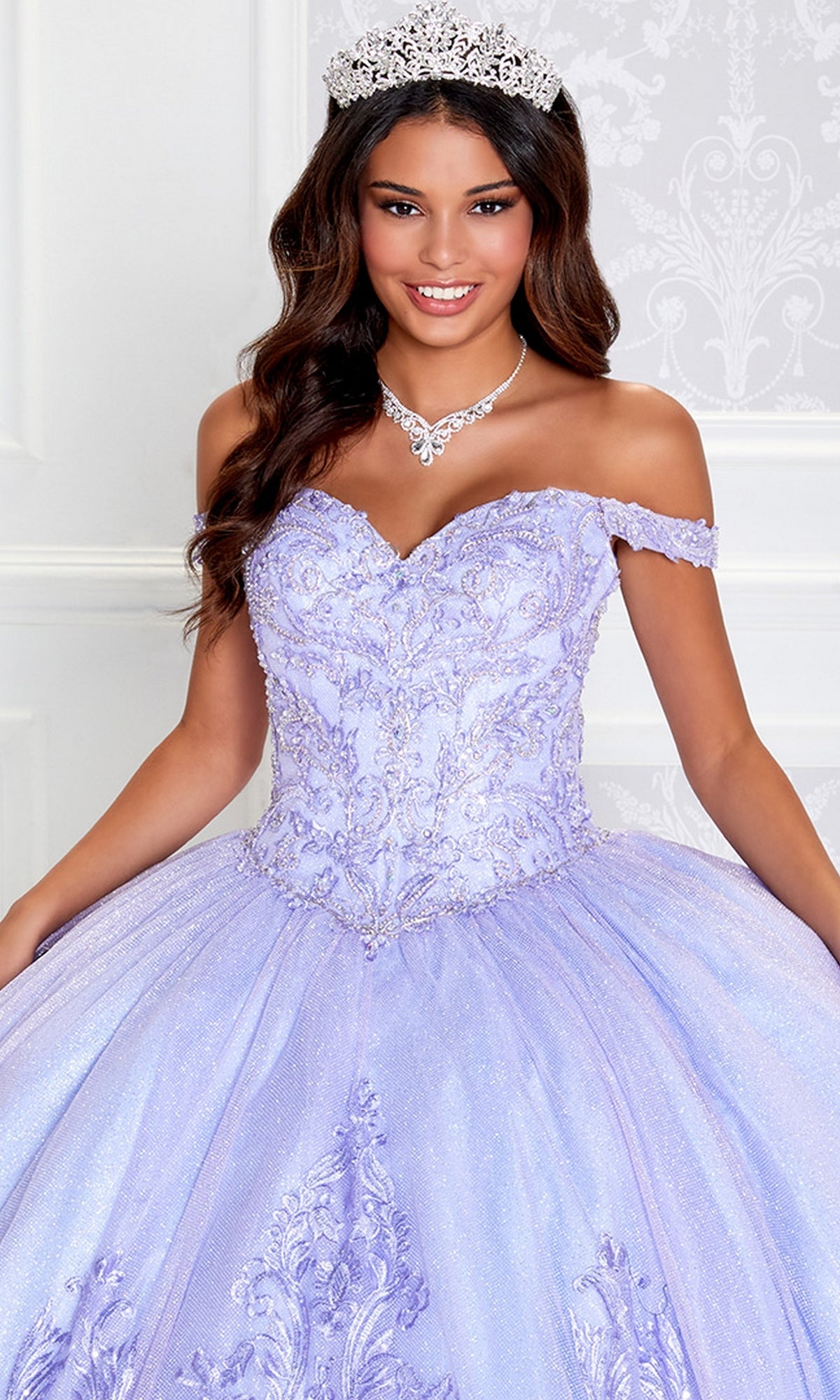 A woman in the PR12264 Princesa Quince Dress with glitter tulle, featuring intricate embroidery and an off-the-shoulder lavender design, poses wearing a tiara and necklace.