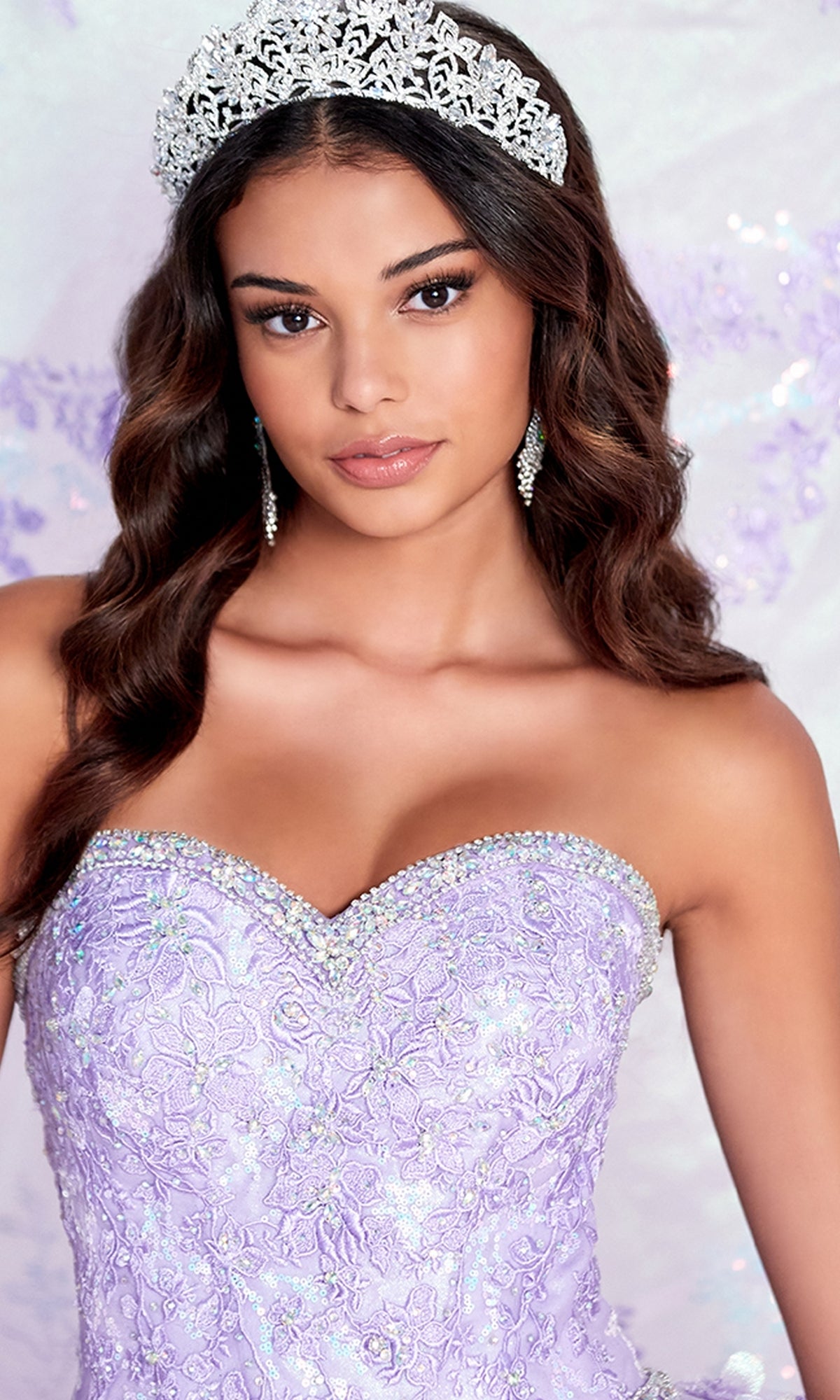 A woman poses in the Strapless Princesa PR12265 Quince Dress with Train, accented by a jeweled tiara and drop earrings, featuring lavender lace and intricate beaded detailing.