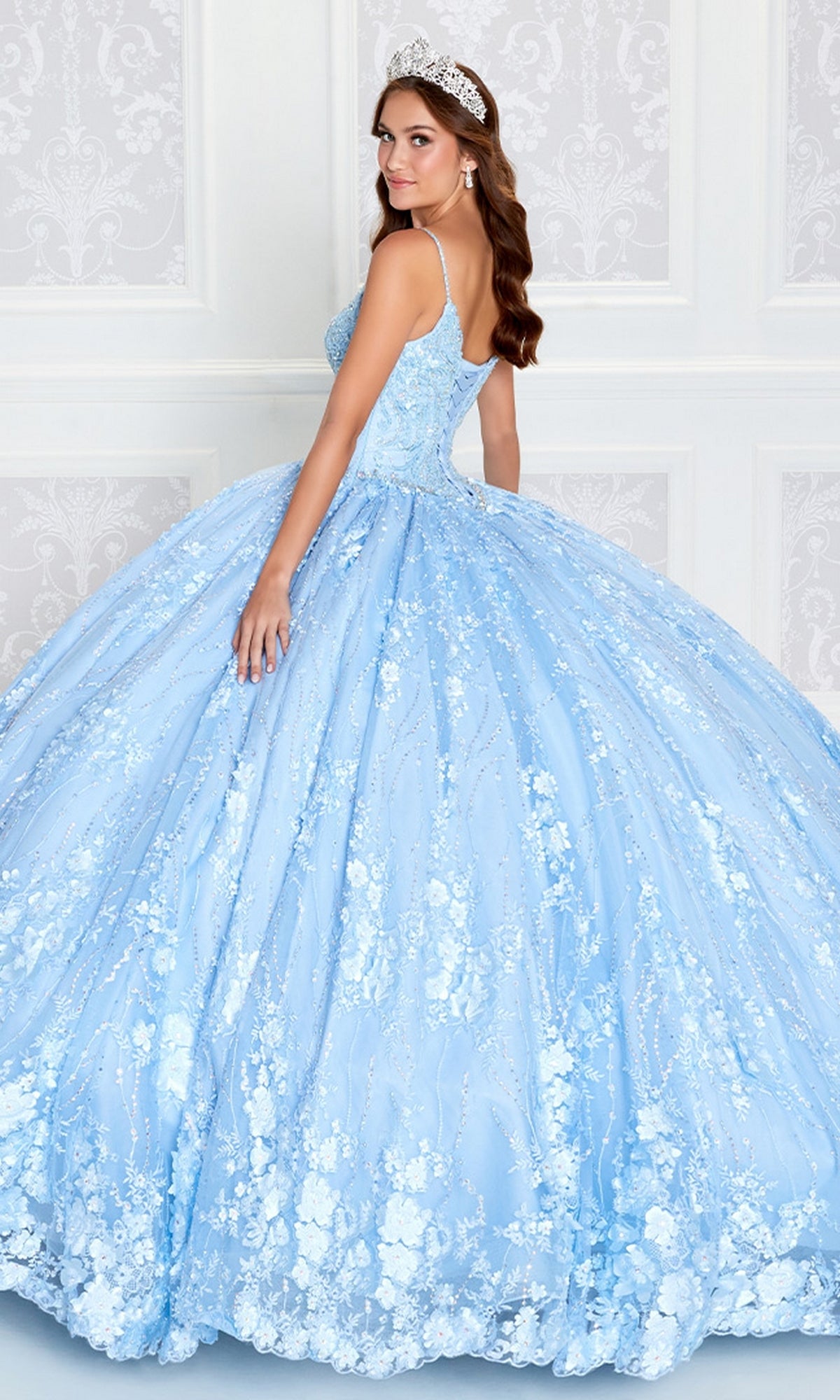 A young woman in a Princesa by Ariana Vara Lace Quince Dress PR12268 and tiara poses before a white patterned wall, glancing over her shoulder.