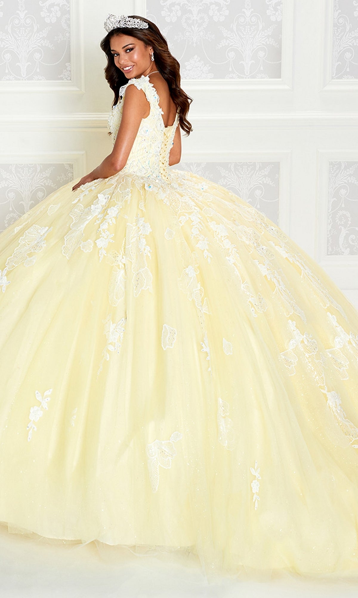 A woman stands in front of a white patterned wall, smiling over her shoulder while wearing the Princesa by Ariana Vara Pastel Quince Dress PR12270 with delicate white lace details and a sparkling tiara.