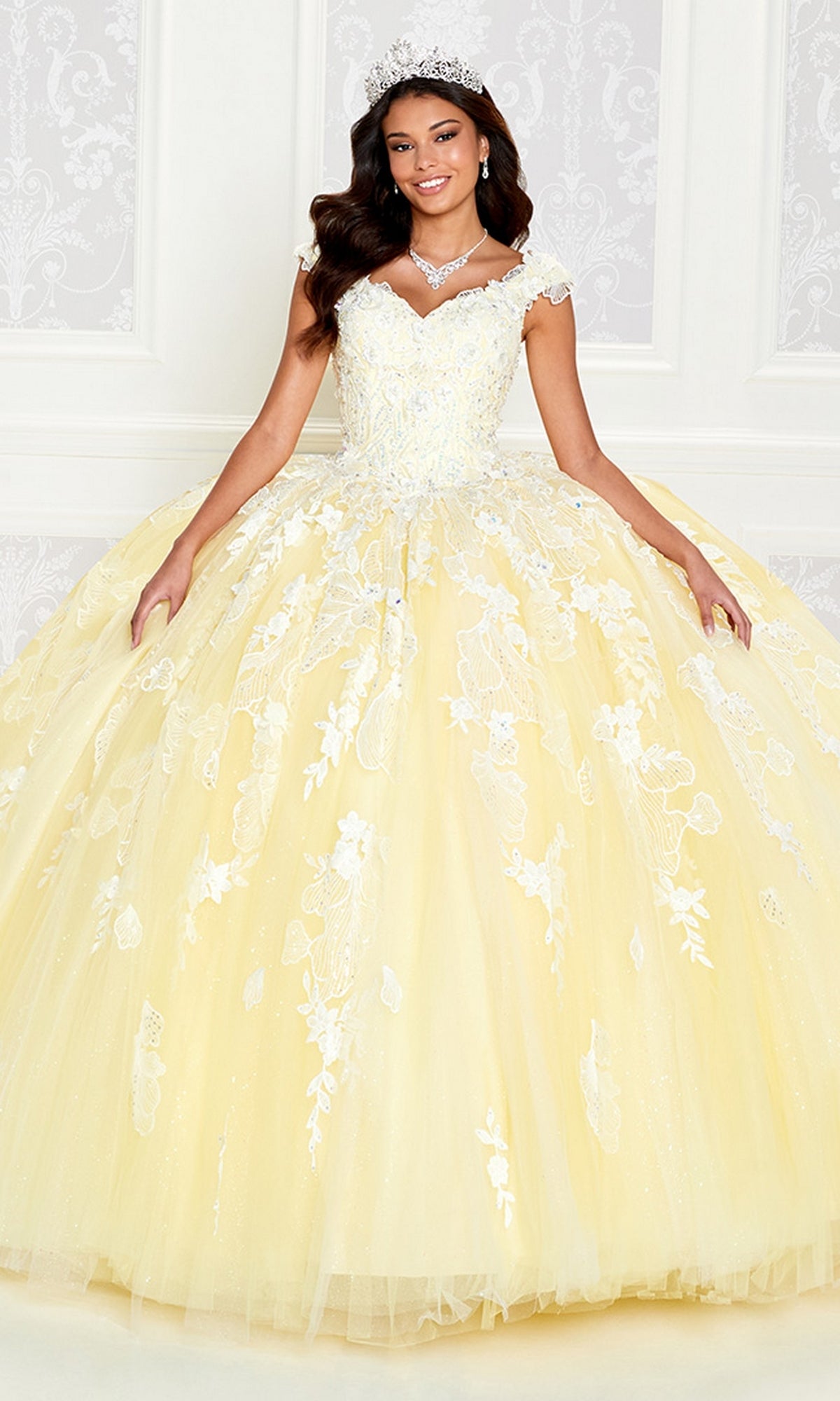 A woman in a yellow off-the-shoulder quinceañera gown with white floral lace, tiara, and necklace stands against a white patterned backdrop, wearing the enchanting Princesa by Ariana Vara Pastel Quince Dress PR12270.