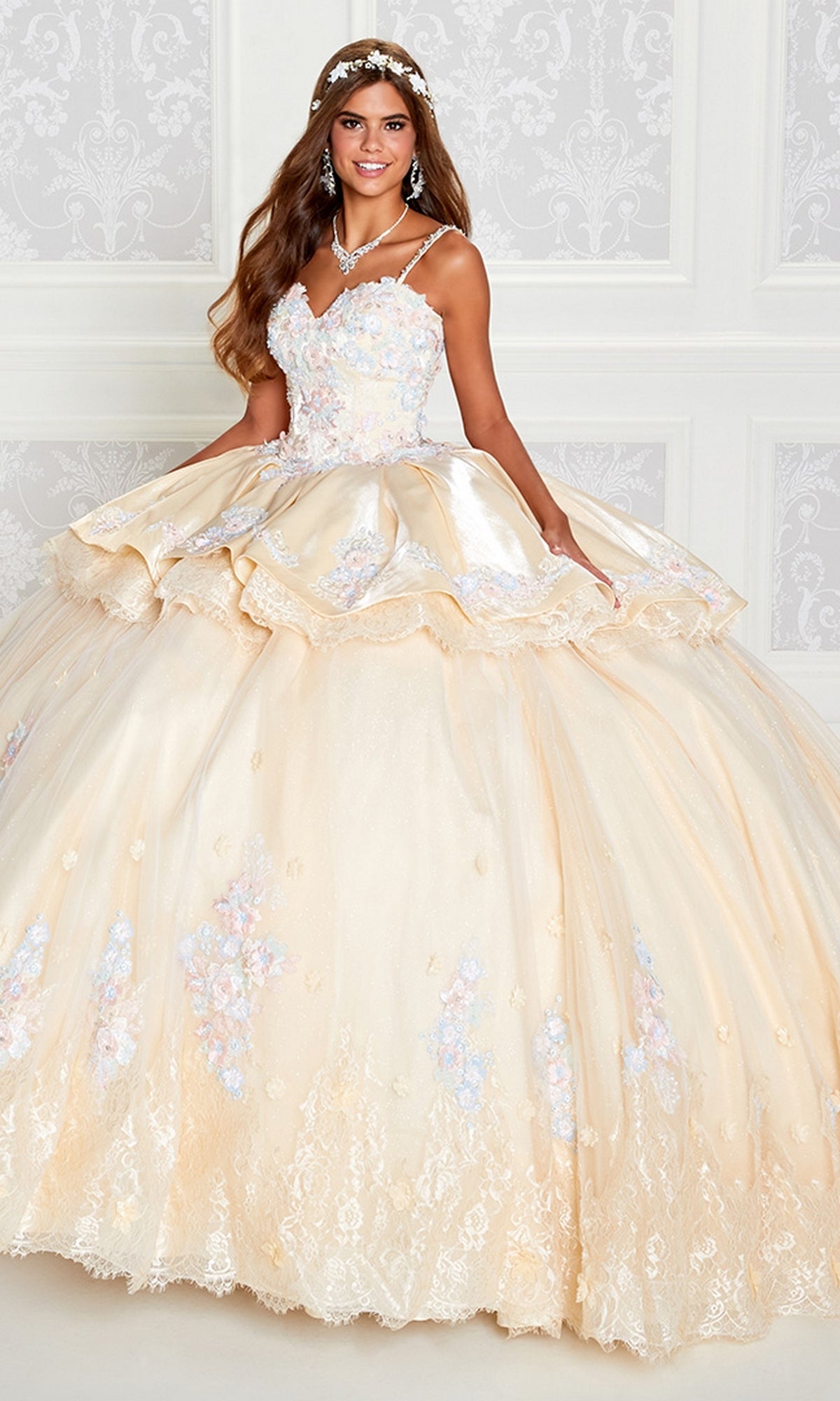 A young woman stands in a formal setting wearing the PR12272 Princesa Quince Dress with Removable Skirt, a cream-colored gown adorned with floral accents, paired with a jeweled necklace and headpiece.