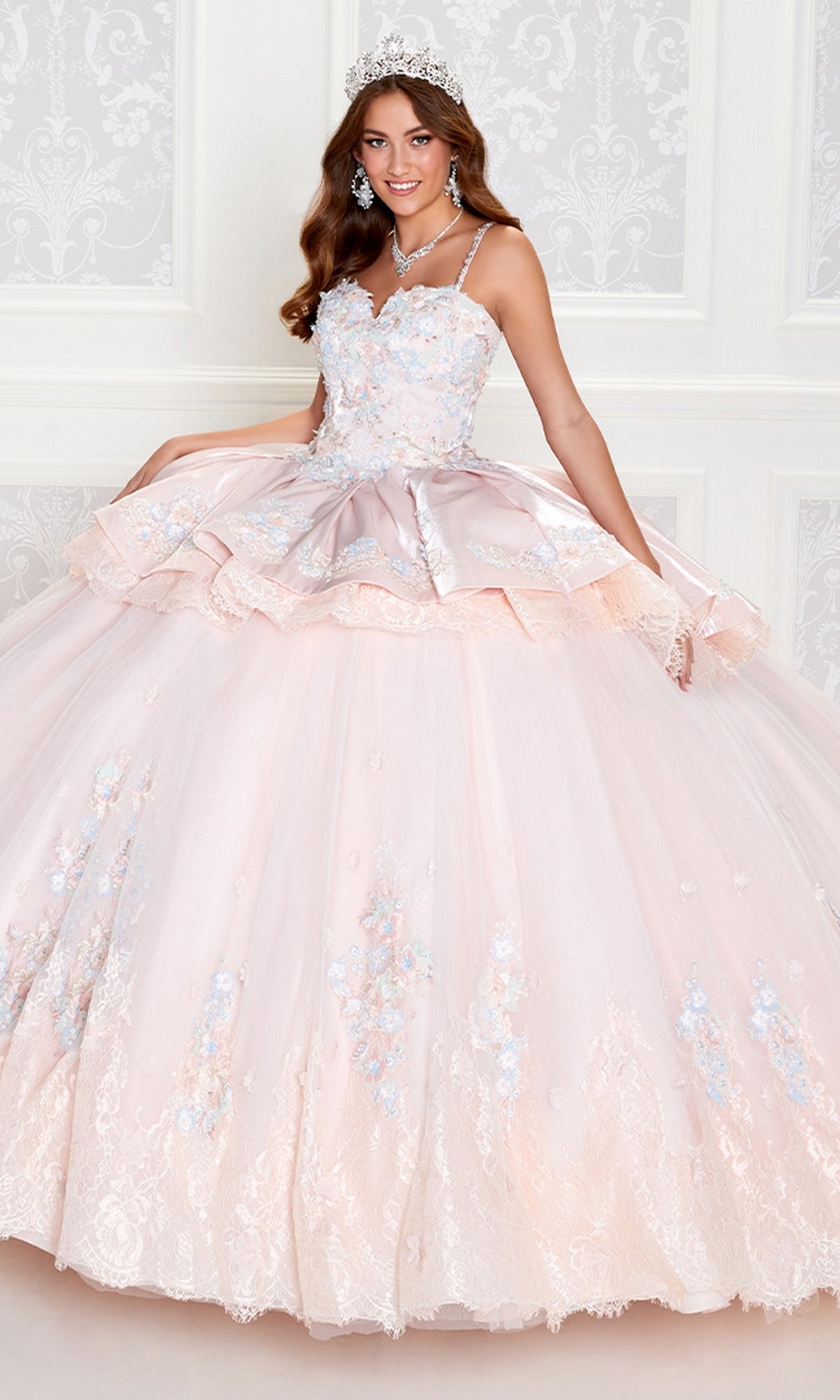 A young woman in the PR12272 Princesa Quince Dress with Removable Skirt, featuring floral embroidery and a jeweled tiara, poses before a white decorative wall.