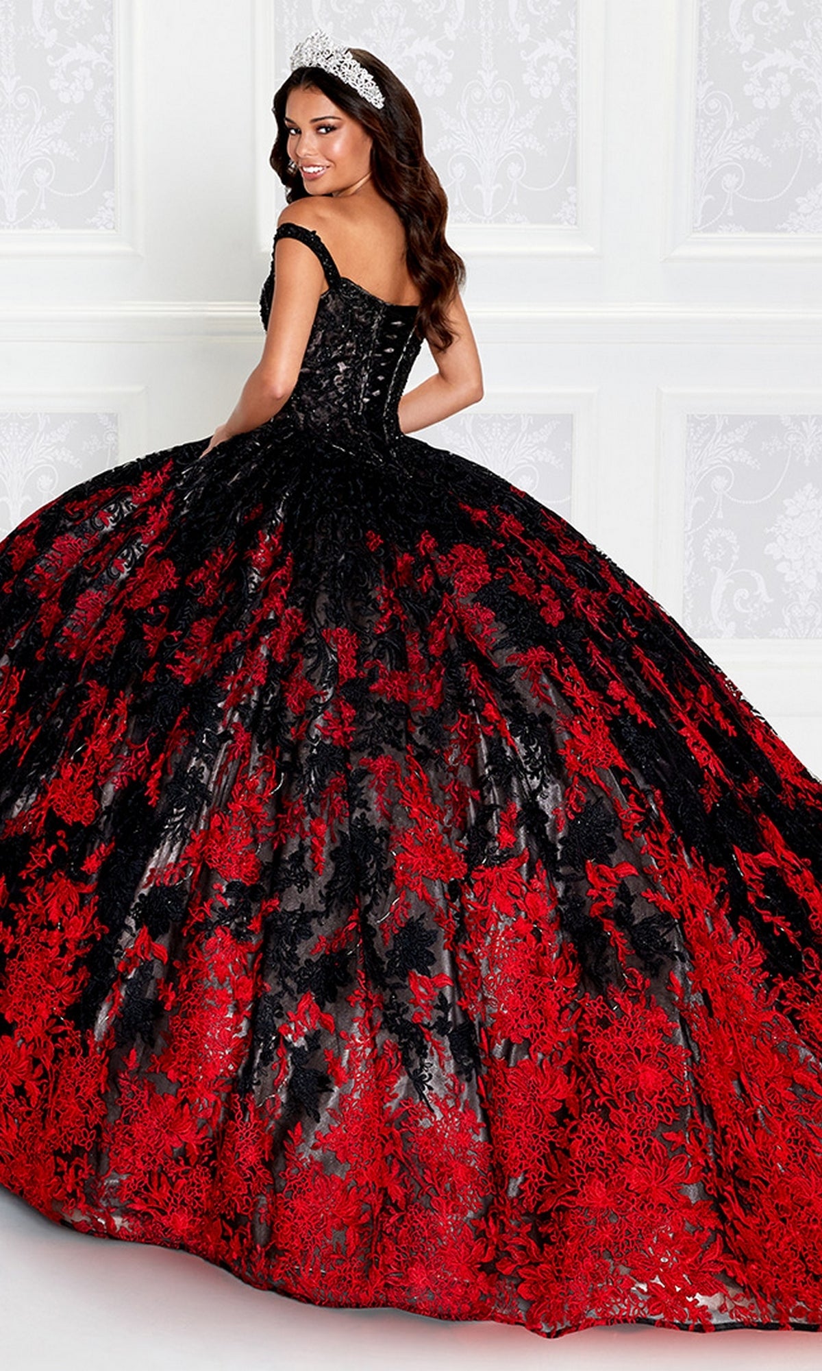 A woman stands before a white patterned wall, wearing the PR12275 Princesa Quinceañera Dress—an off-shoulder gown with a black bodice and a full skirt adorned with black and red embroidered lace floral designs.