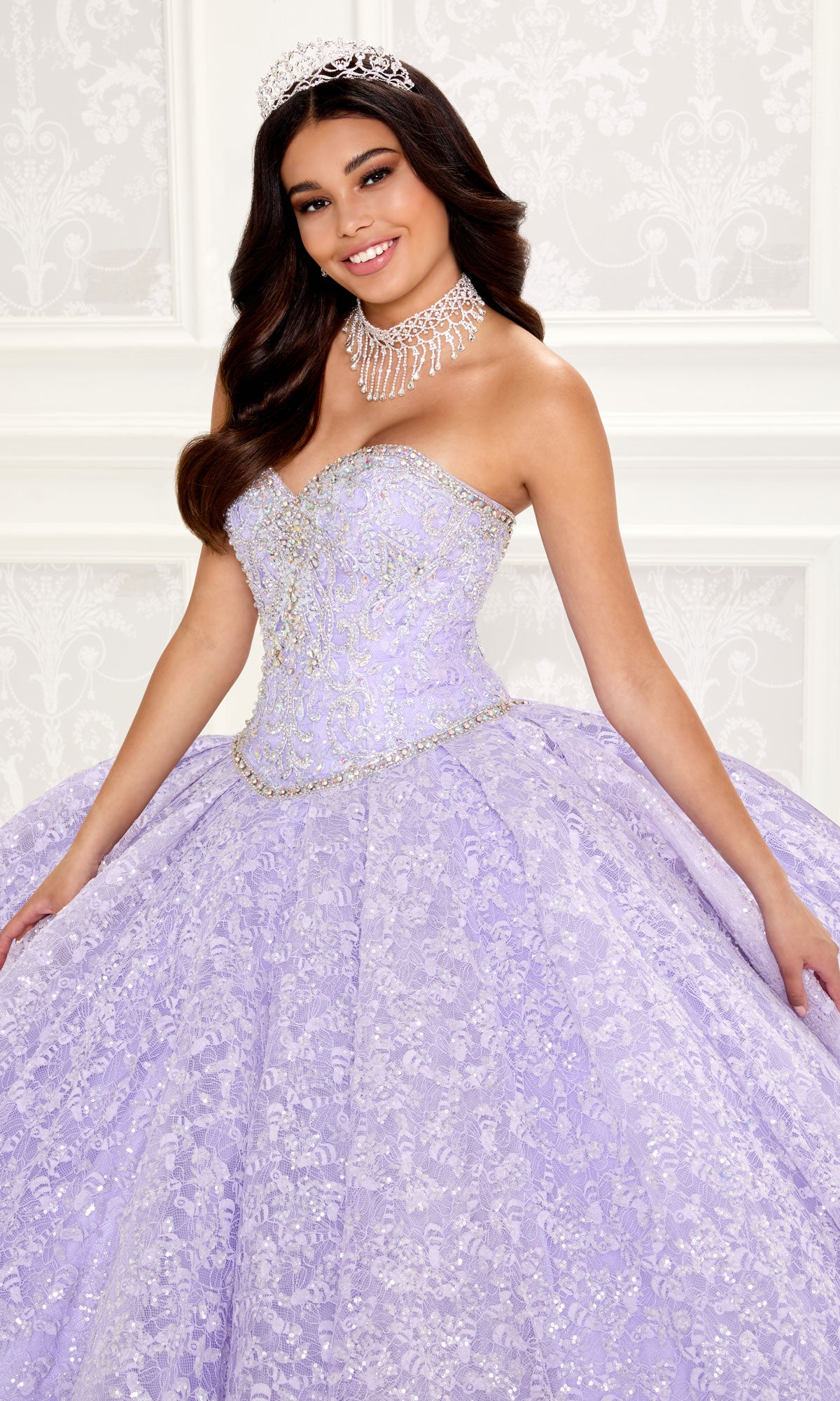 A young woman smiles at the camera indoors, wearing a jeweled tiara and the Strapless Quinceanera Dress: Princesa PR22022 in ornate lavender.