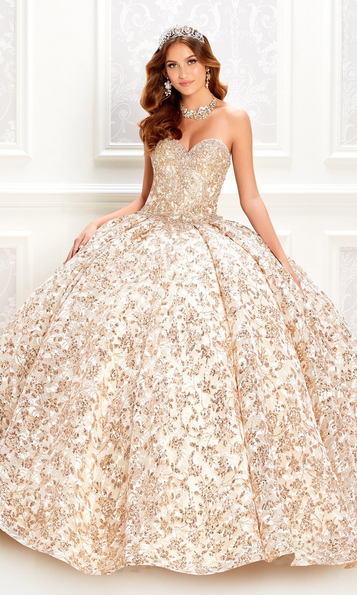 A woman models the Strapless Quinceanera Dress: Princesa PR22022 in gold and white, featuring a full skirt, tiara, and statement necklace as she poses against an ornate white background.