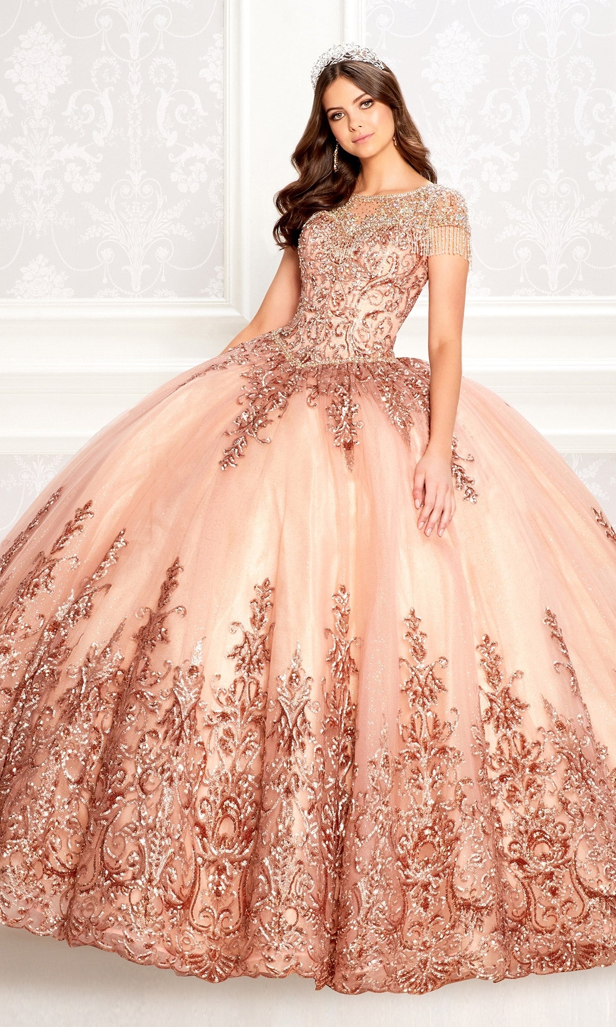 A woman wearing the Princesa PR22026 short-sleeve quinceanera dress with a beaded bodice and gold embroidery, paired with a tiara, stands before an ornate white background.