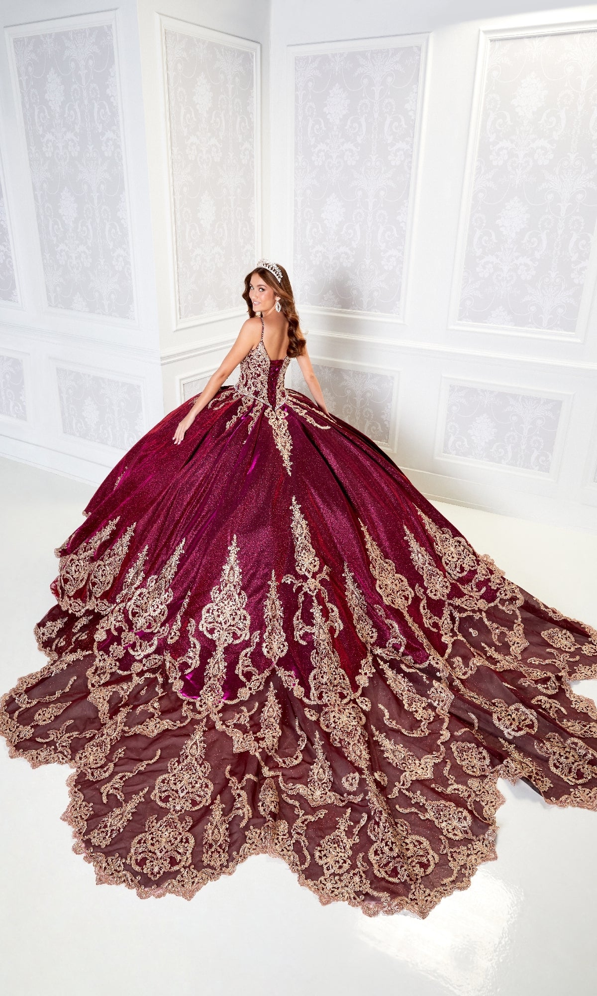 A woman in the Quinceanera Dress PR22141 By Princesa, featuring gold embroidery and a corset back, poses with a tiara in an ornate white room, exuding elegance in her luxurious burgundy ball gown.
