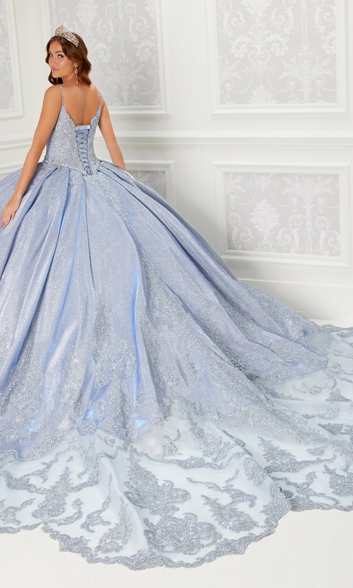 A woman in the Princesa Quinceanera Dress PR22141, featuring a blue ball gown, corset back, long ornate train, and silver embroidery, stands in an elegant white-decorated room, looking over her shoulder.