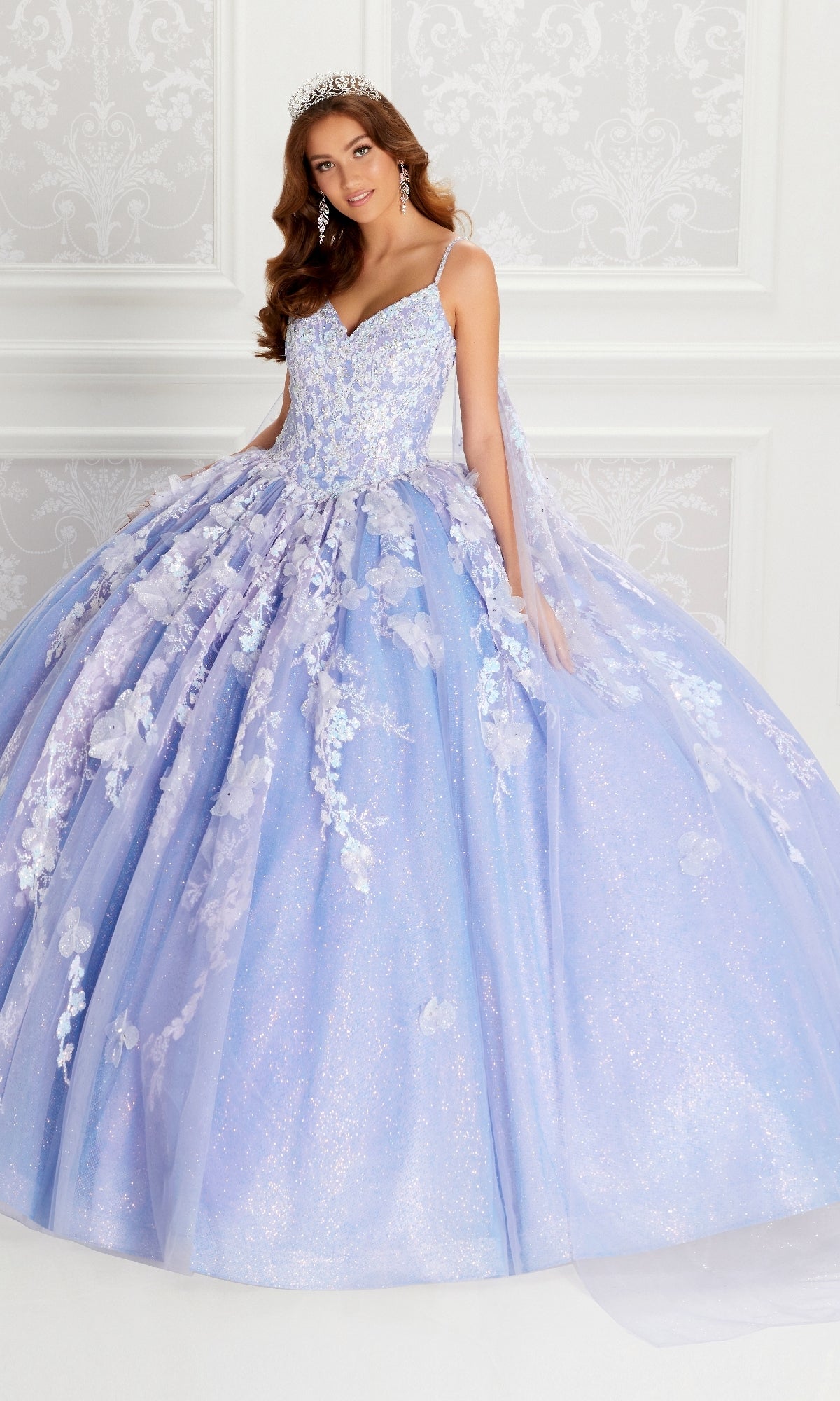 A young woman in the Quinceanera Dress PR22143 By Princesa, featuring sparkling lavender fabric, floral appliqués, a beaded corset bodice, and a tiara, stands against a white ornate background.