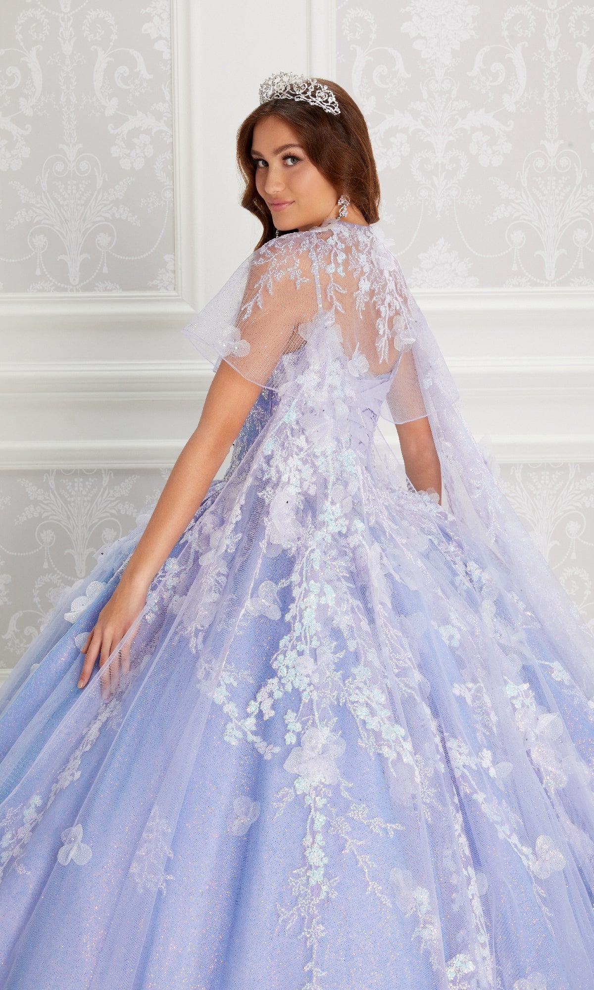 A young woman wearing the Quinceanera Dress PR22143 By Princesa, featuring a sparkly lavender hue, beaded corset bodice, floral lace details, and a tiara, poses before a patterned wallpaper backdrop.