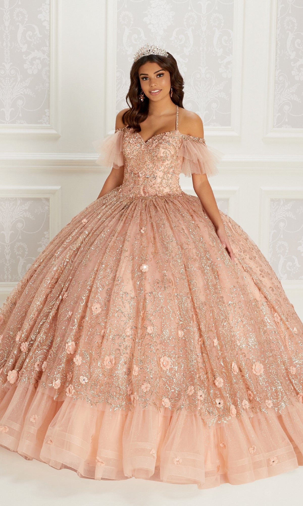 A young woman wears the Quinceanera Dress PR22144 by Princesa—a peach ball gown with off-the-shoulder sleeves, corset back, and sparkling floral details—paired with a jeweled tiara, standing against an ornate white background.