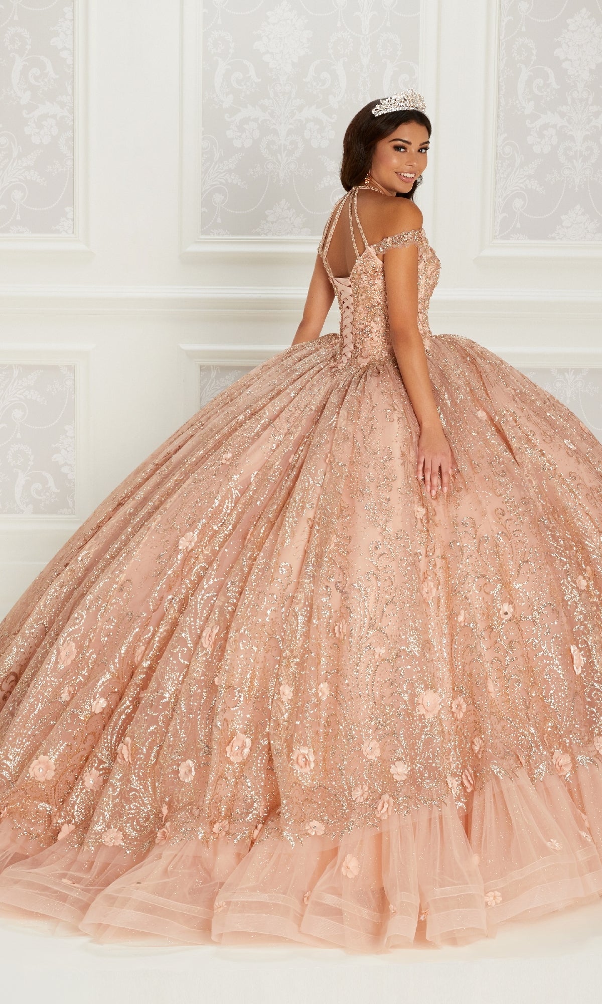 A woman in the Quinceanera Dress PR22144 By Princesa, featuring peach-colored glitter, intricate beading, and embroidery stands in an elegant room, wearing a tiara and looking over her shoulder.