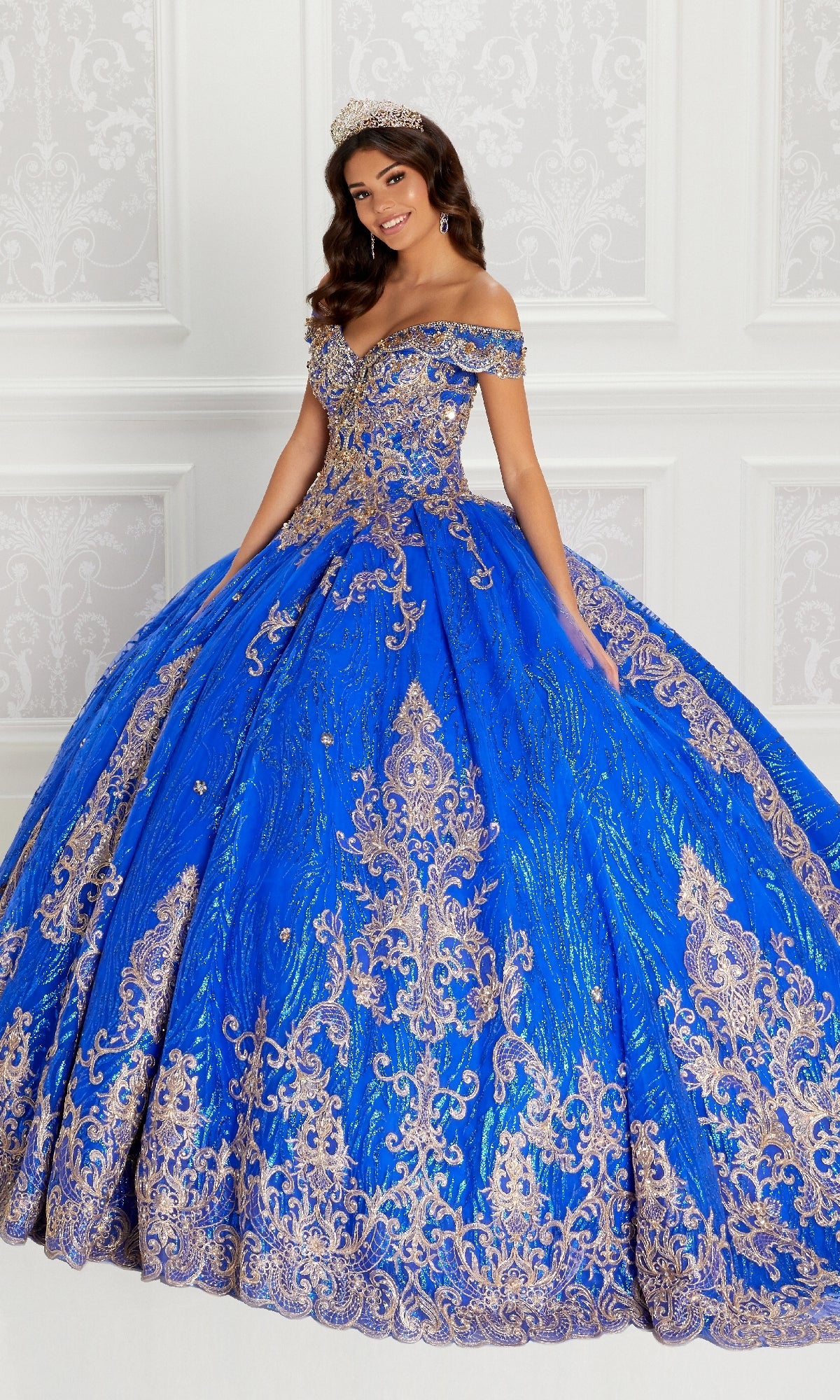 A woman stands gracefully in front of a white paneled wall wearing the Quinceanera Dress PR22148 By Princesa, an ornate off-shoulder royal blue gown with gold lace embroidery, beaded accents, and a sparkling jeweled tiara.