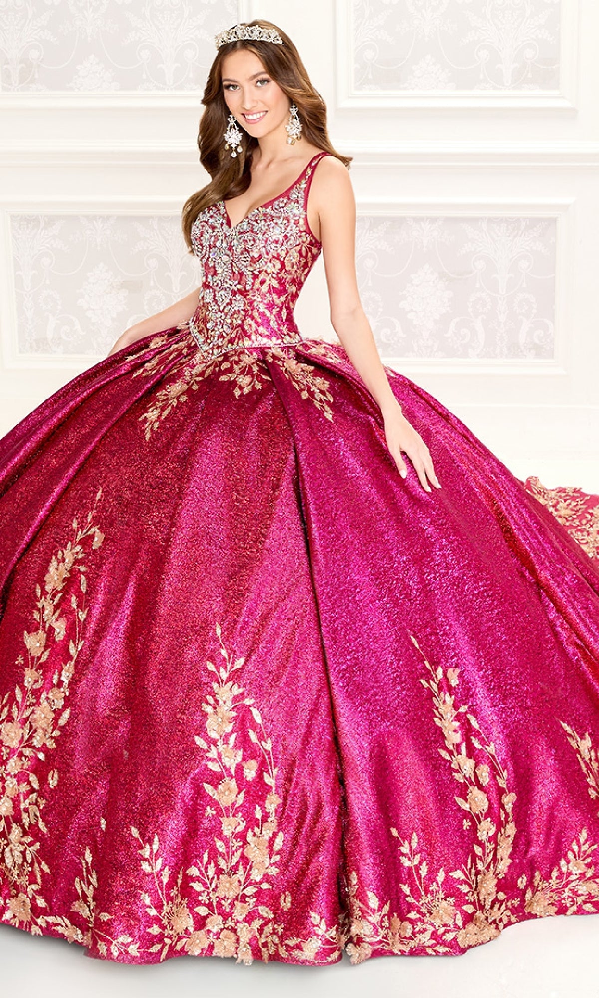 A woman in the Quinceanera Dress PR30081 By Princesa—sparkling magenta metallic jacquard with gold floral embroidery—wears matching earrings and a tiara as she stands in a decorated room.