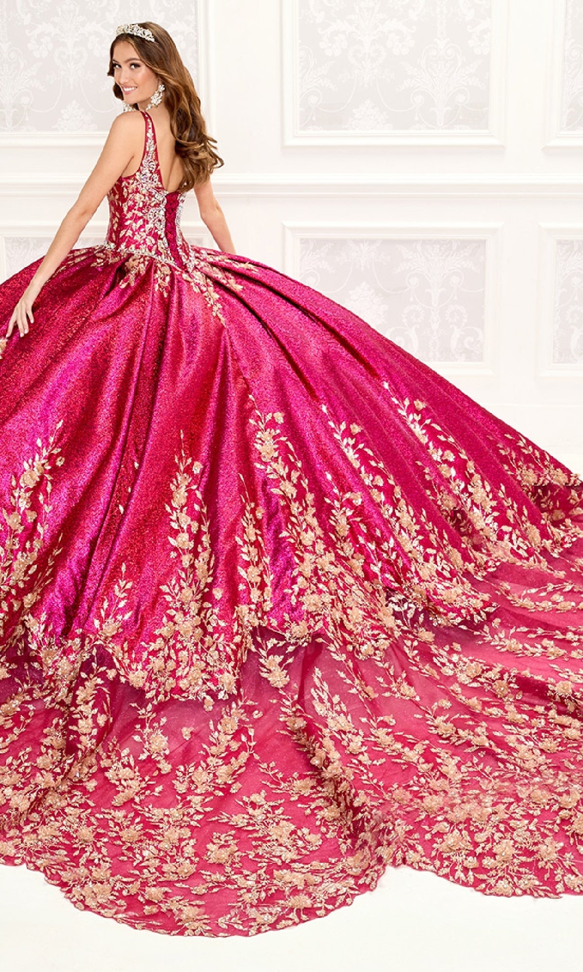 A woman in the Quinceanera Dress PR30081 By Princesa, a vibrant pink metallic jacquard gown with gold floral embroidery and a long train, stands in an ornate white room.