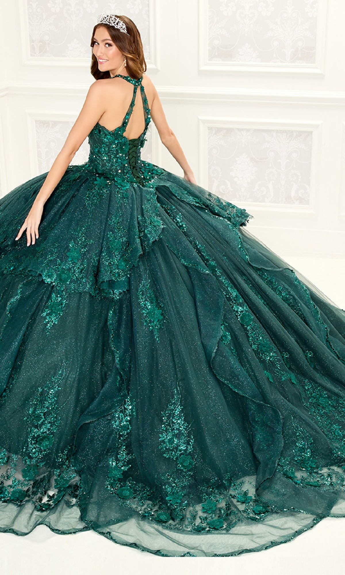 A woman wearing the Quinceanera Dress PR30082 By Princesa, featuring green embroidery with lace and sequin details, stands in a white room, smiling over her shoulder.