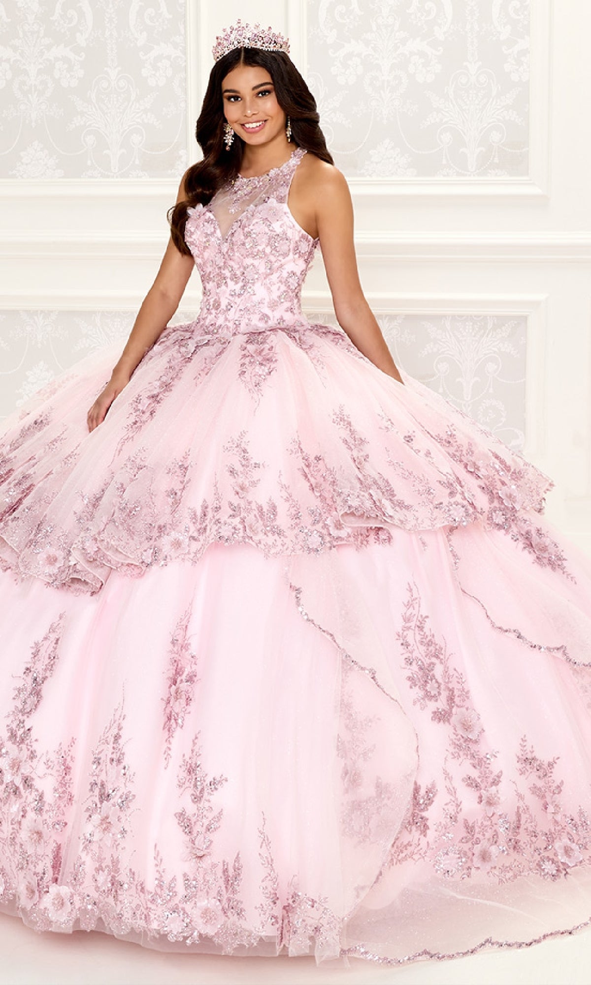 A young woman wearing the Quinceanera Dress PR30082 By Princesa with floral embroidery and a matching tiara stands in an elegant, white-decorated room.