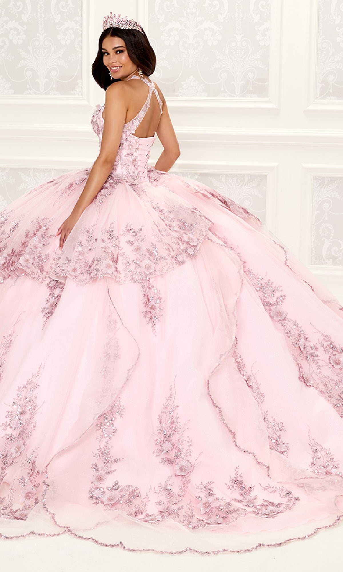 A young woman in the Quinceanera Dress PR30082 By Princesa, featuring a pink hue and lace detailing with an illusion neckline, stands in a white ornate room, wearing a tiara and smiling over her shoulder.