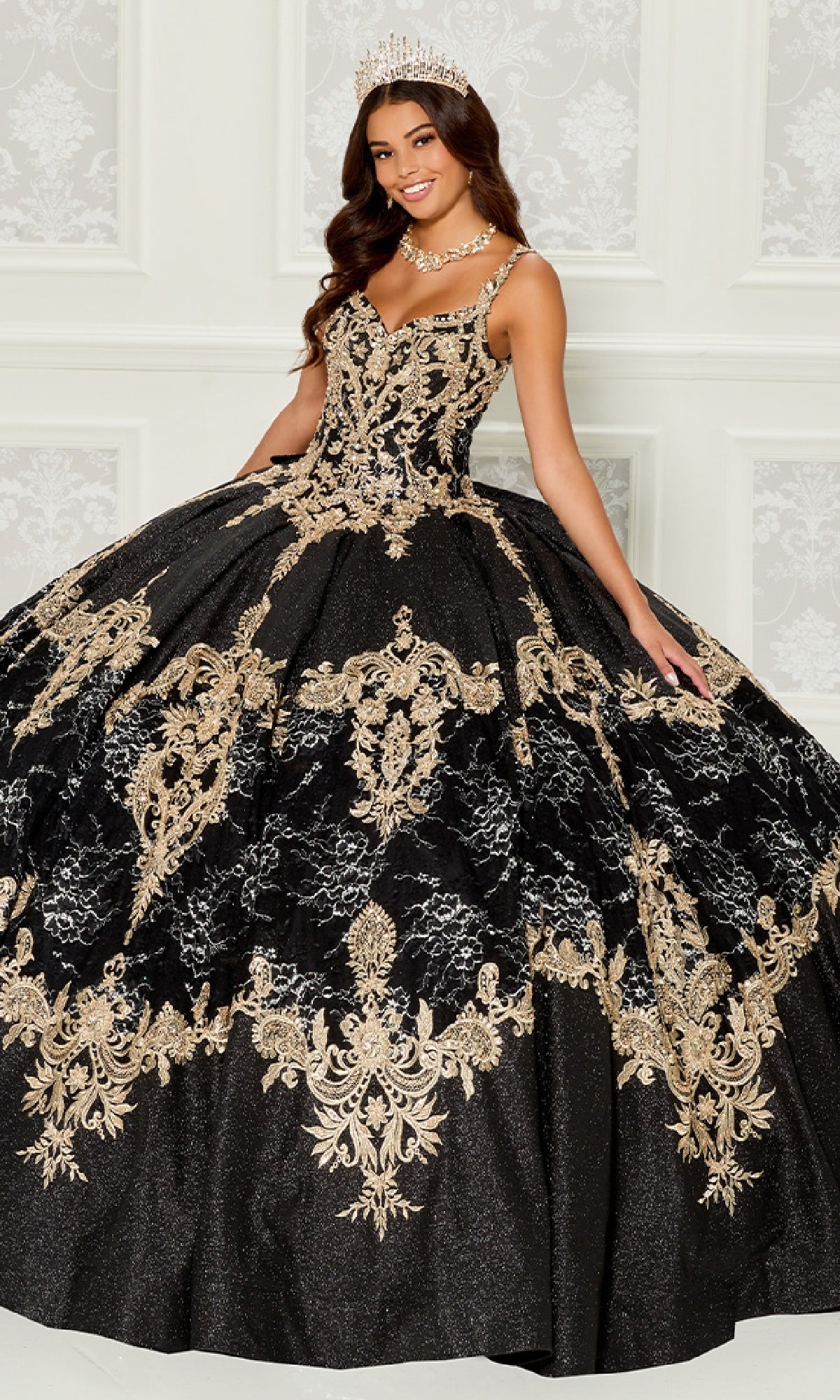 A woman smiles indoors, wearing the Quinceanera Dress PR30085 By Princesa—a black gown with gold embroidery and beading—holding out part of her skirt and accessorized with a tiara.