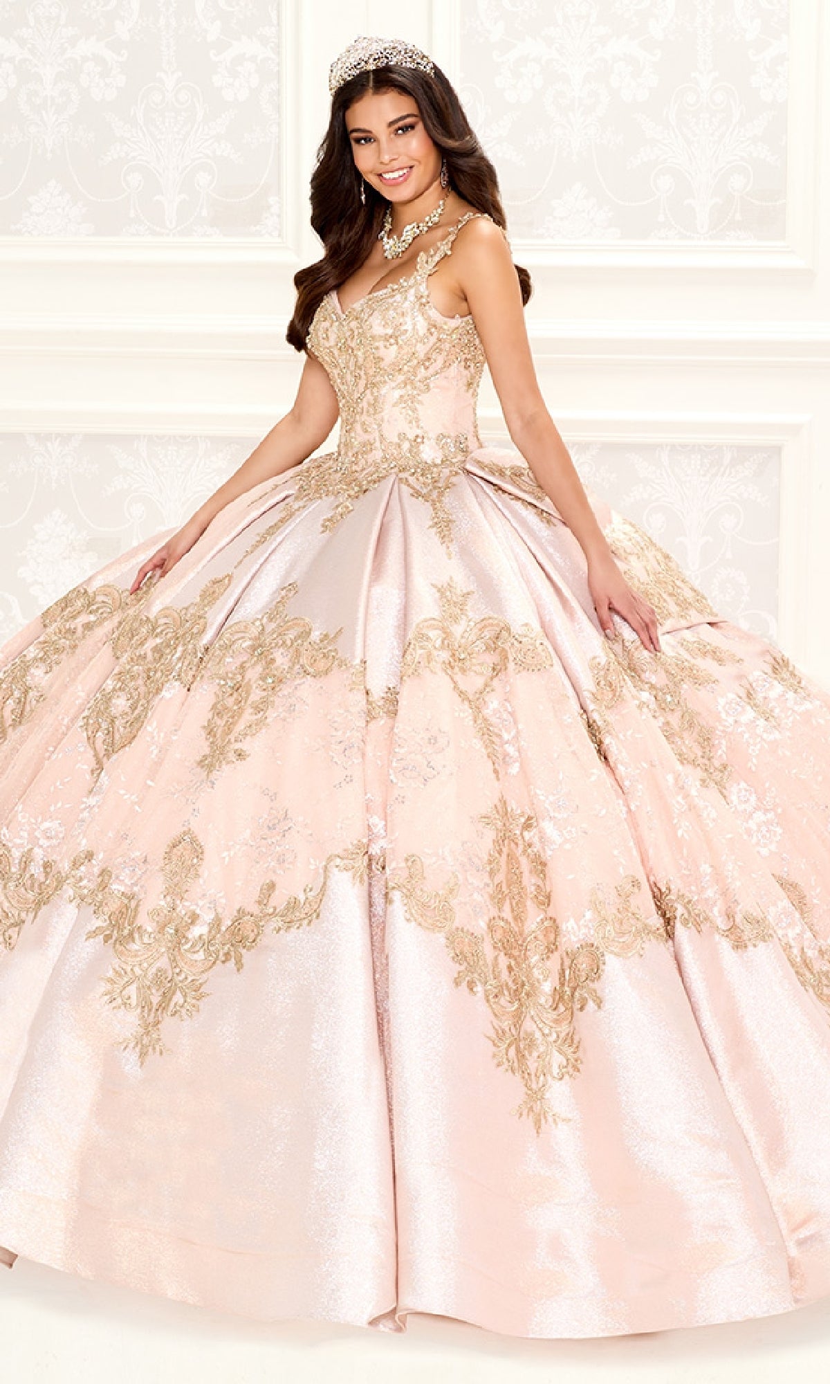 A young woman stands in a formal setting wearing the Quinceanera Dress PR30085 By Princesa, featuring pink and gold metallic embroidered lace, elegant beading, a tiara, and a necklace.