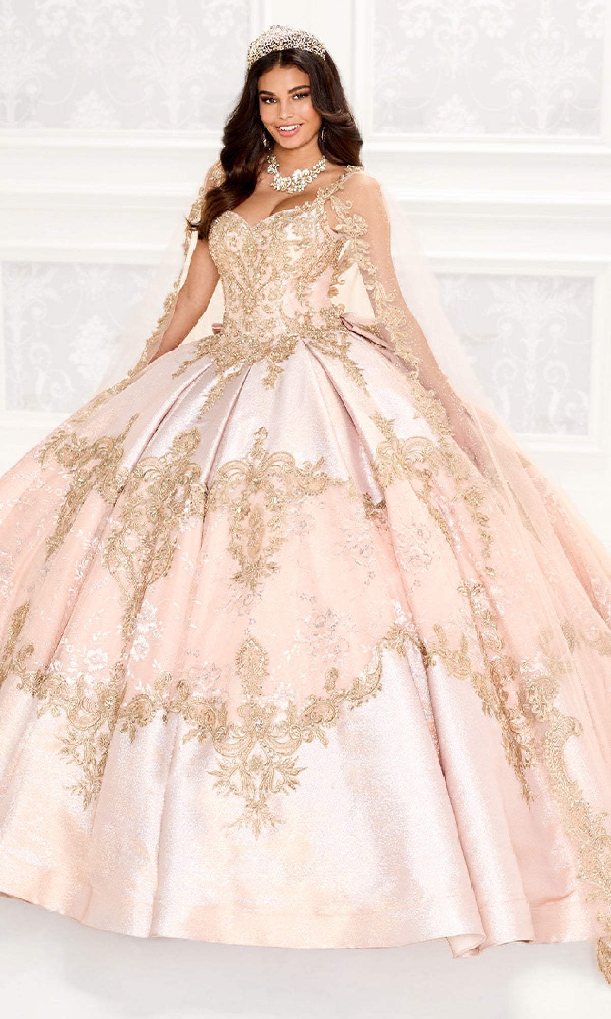 A woman smiles in front of a white patterned background, wearing the Quinceanera Dress PR30085 By Princesa—an ornate pink gown with gold beading details and a jeweled tiara.