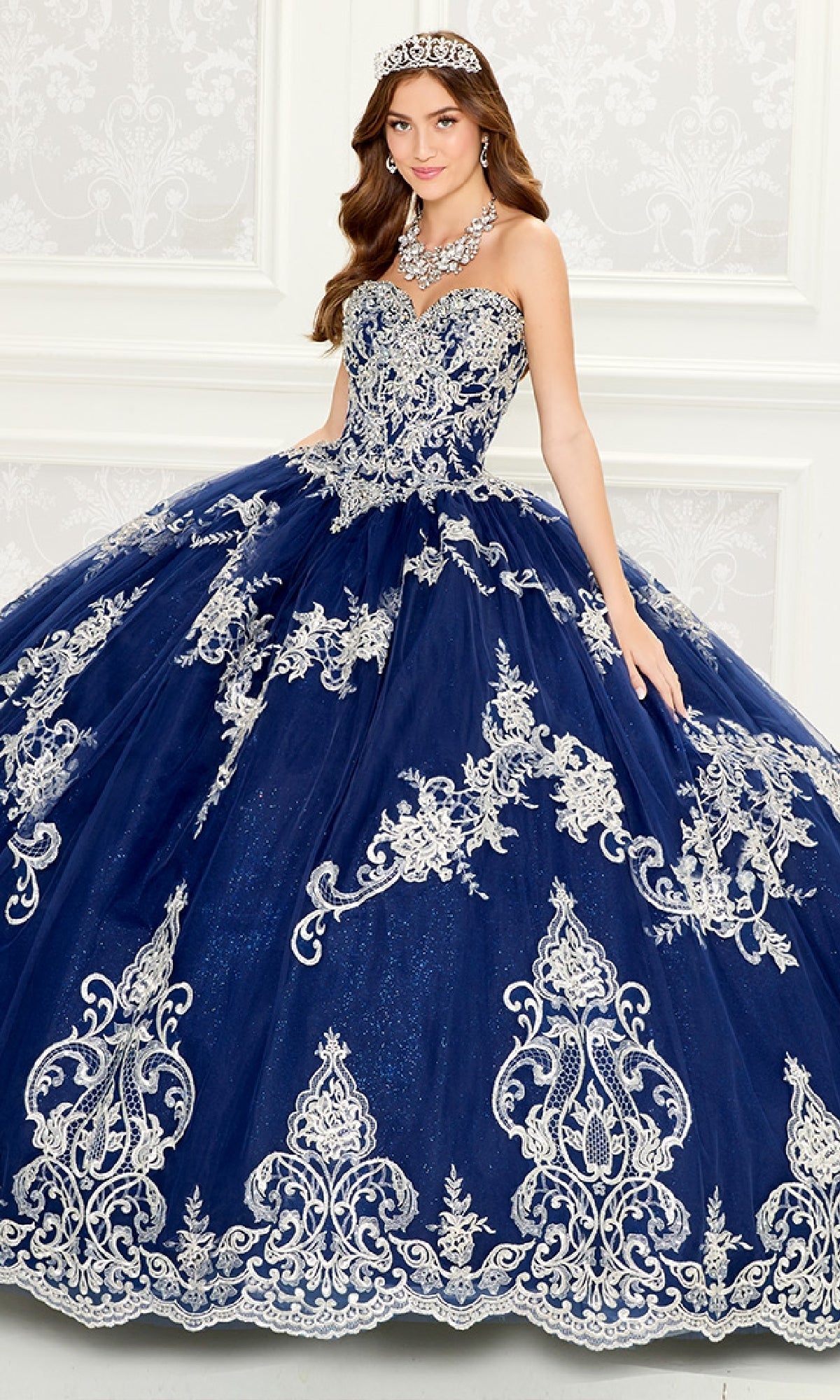 A woman poses indoors against a white decorative wall wearing the Quinceanera Dress PR30086 By Princesa, a navy blue ball gown with silver embroidery, a sheer cape, and accessorized with a jeweled tiara and matching necklace.