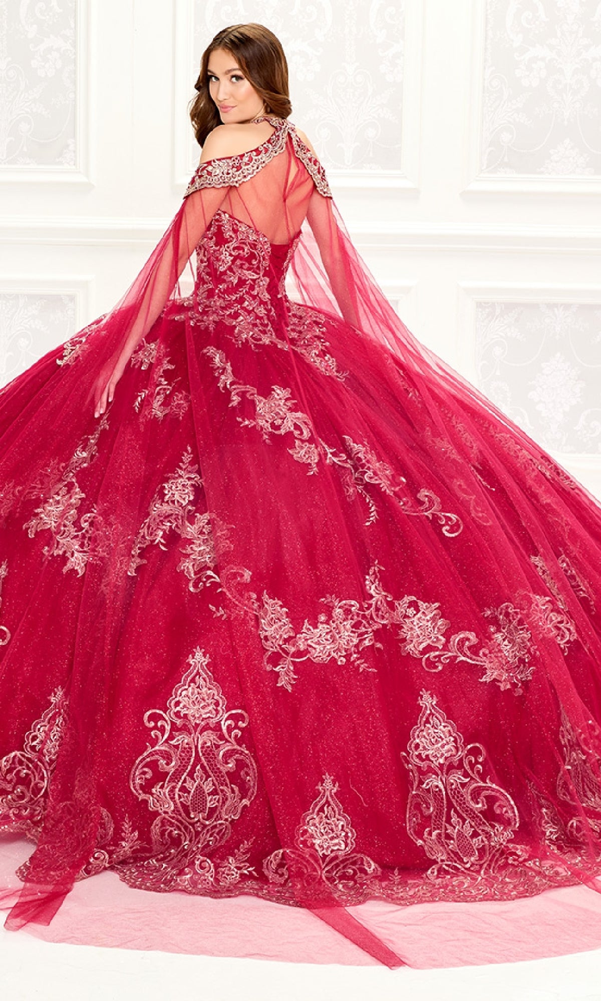A woman wearing the Quinceanera Dress PR30086 By Princesa—with elaborate red fabric, silver embroidery, and tulle details—stands in an ornate white room, facing slightly away from the camera.