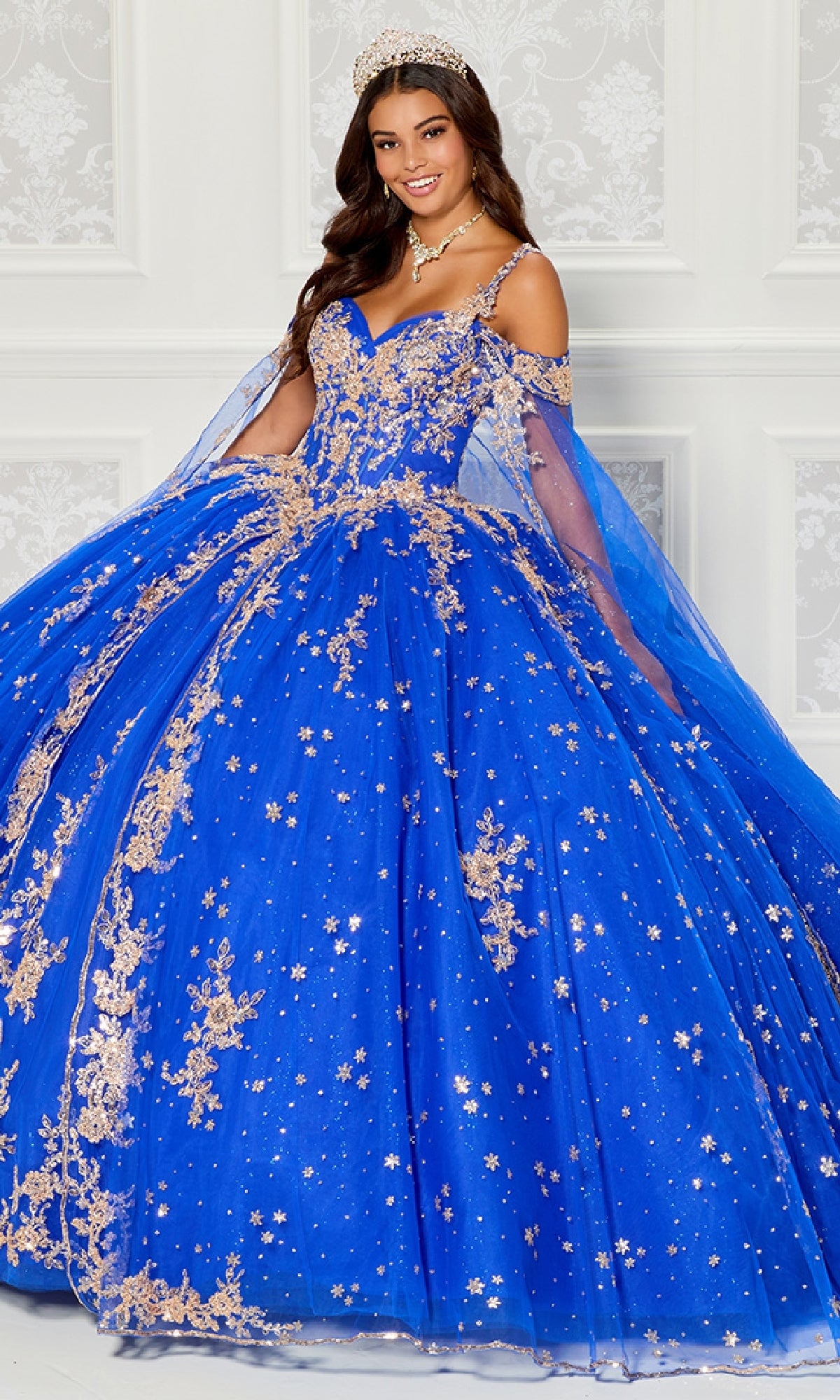 A young woman wears the Quinceanera Dress PR30087 By Princesa, a bright blue gown with gold embellishments and a corset bodice, paired with a tiara, standing before a white decorative wall.