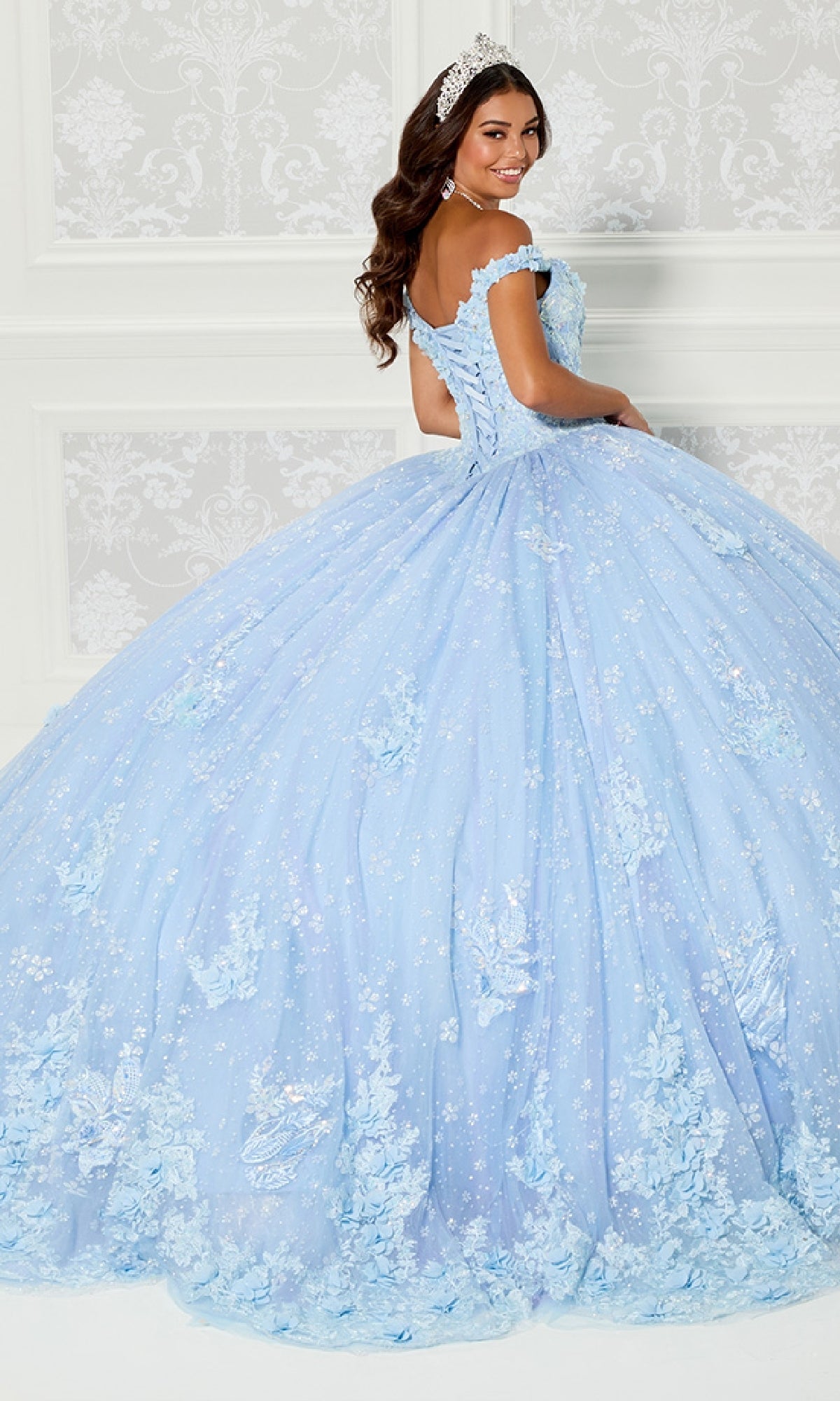 A woman smiles in an elegant room wearing the Quinceanera Dress PR30111 By Princesa—a light blue, off-the-shoulder gown with floral details, lace embroidery, and a sparkling tiara, standing with her back to the camera.