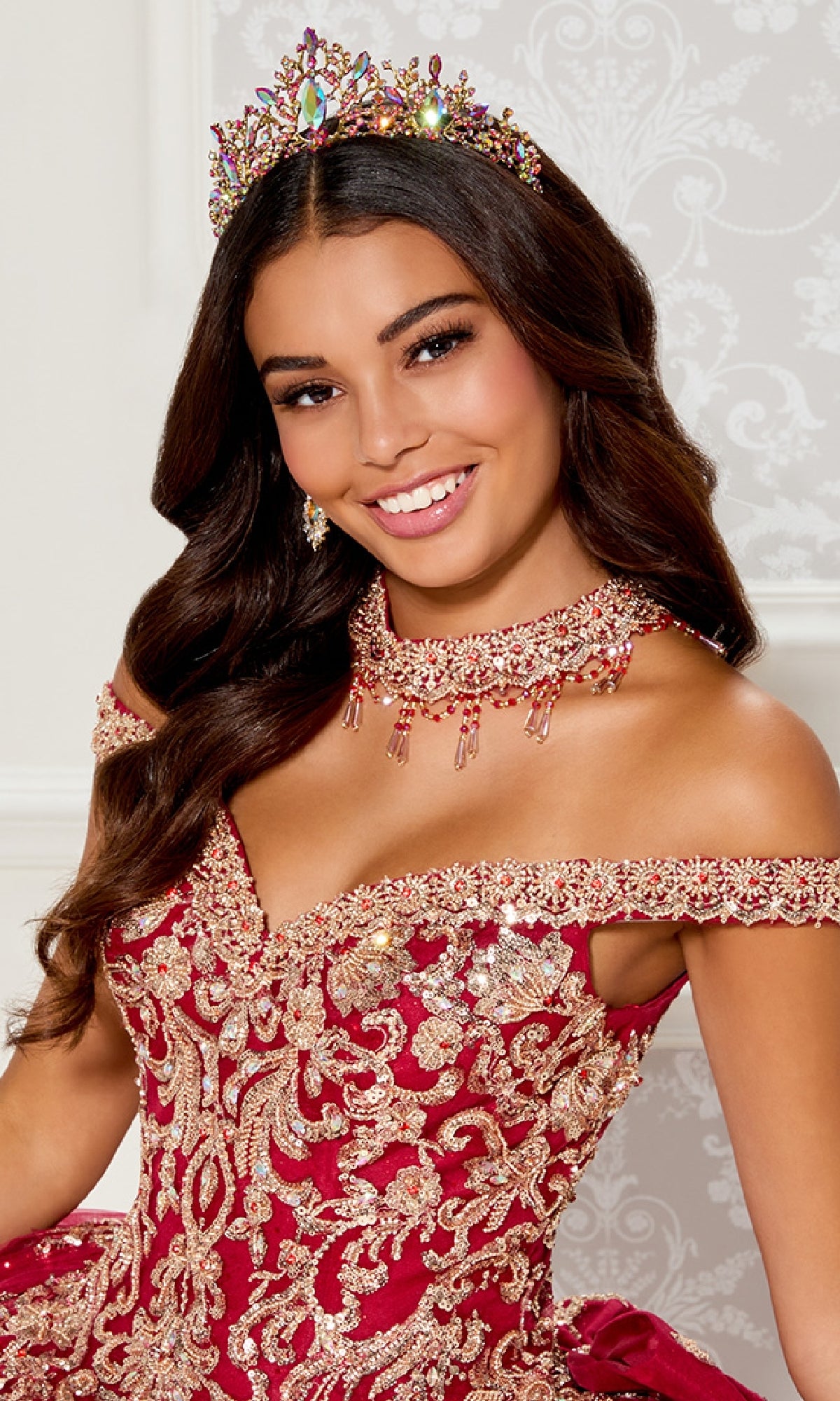 A woman with long, wavy brown hair wears a jeweled crown and the Quinceanera Dress PR30112 By Princesa, featuring a red and gold off-the-shoulder design, as she smiles against a light patterned background.