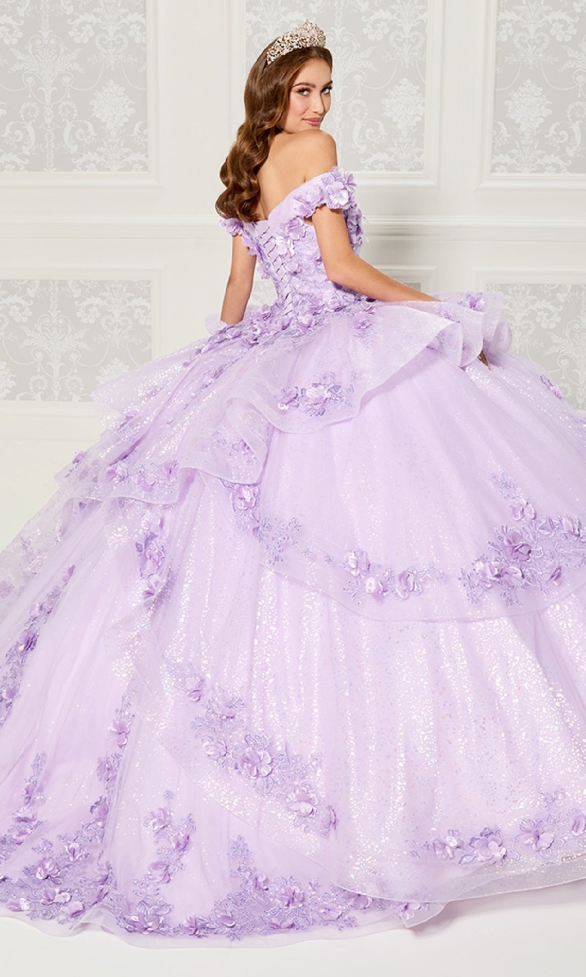 A woman in the Quinceanera Dress PR30113 By Princesa, featuring floral appliqué and a tiara, stands before an ornate white wall, her elegant lavender off-the-shoulder gown radiating grace.