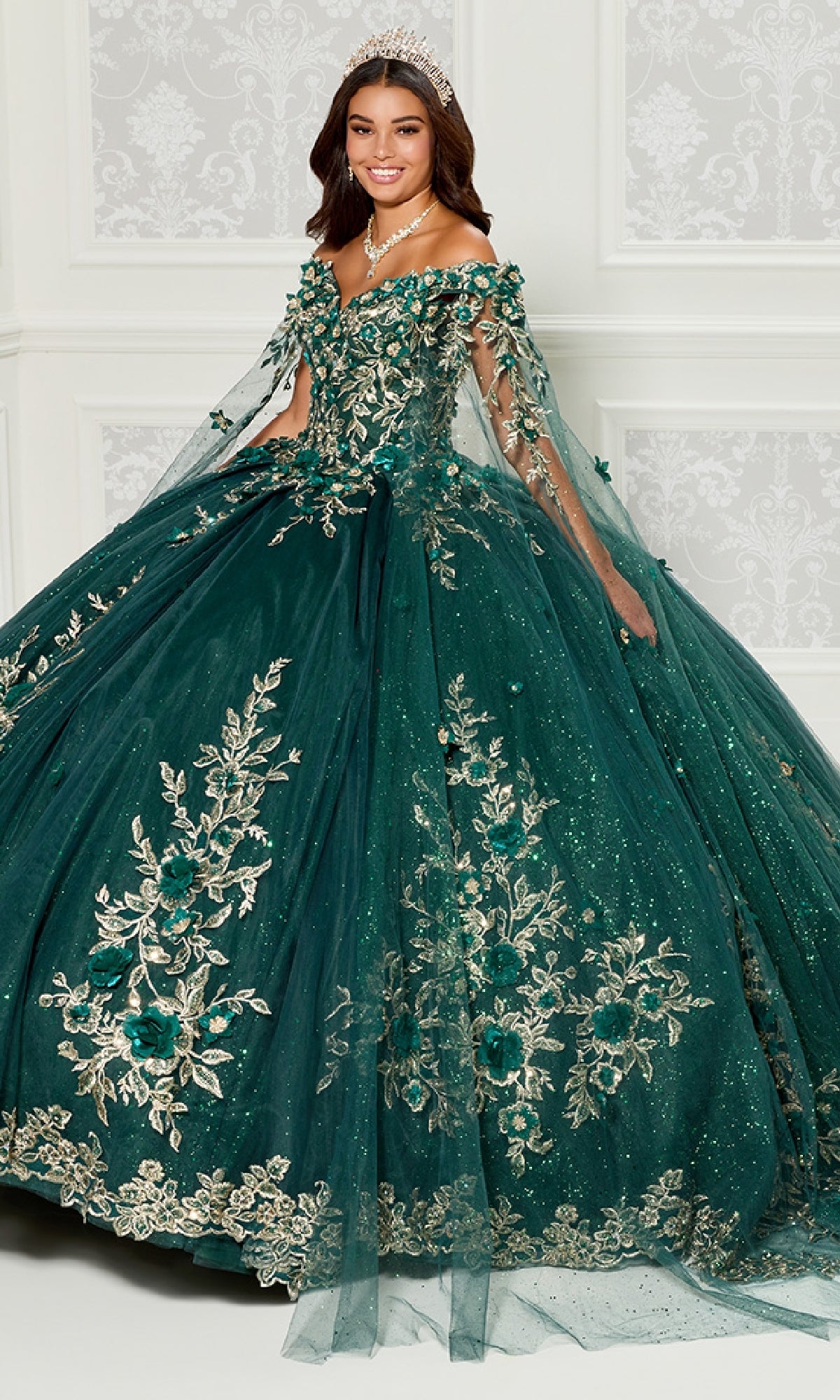 A woman in the Quinceanera Dress PR30114 by Princesa, featuring gold 3D floral appliques and an off-the-shoulder design, wears a tiara while standing in an elegant room with white paneled walls.