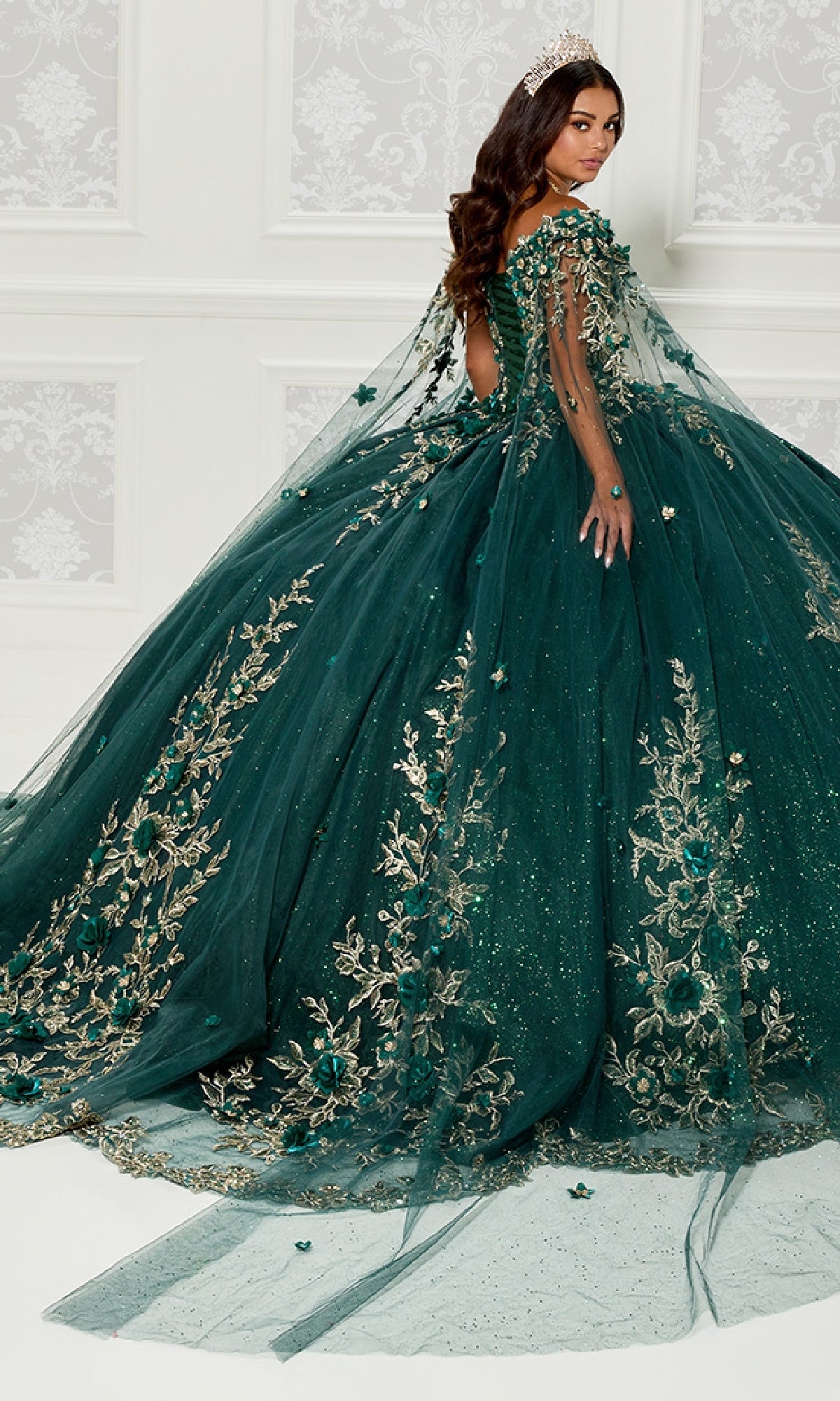A woman wearing the Quinceanera Dress PR30114 By Princesa, featuring gold embroidery and a long train, stands before a white wall with a tiara, glancing over her shoulder.