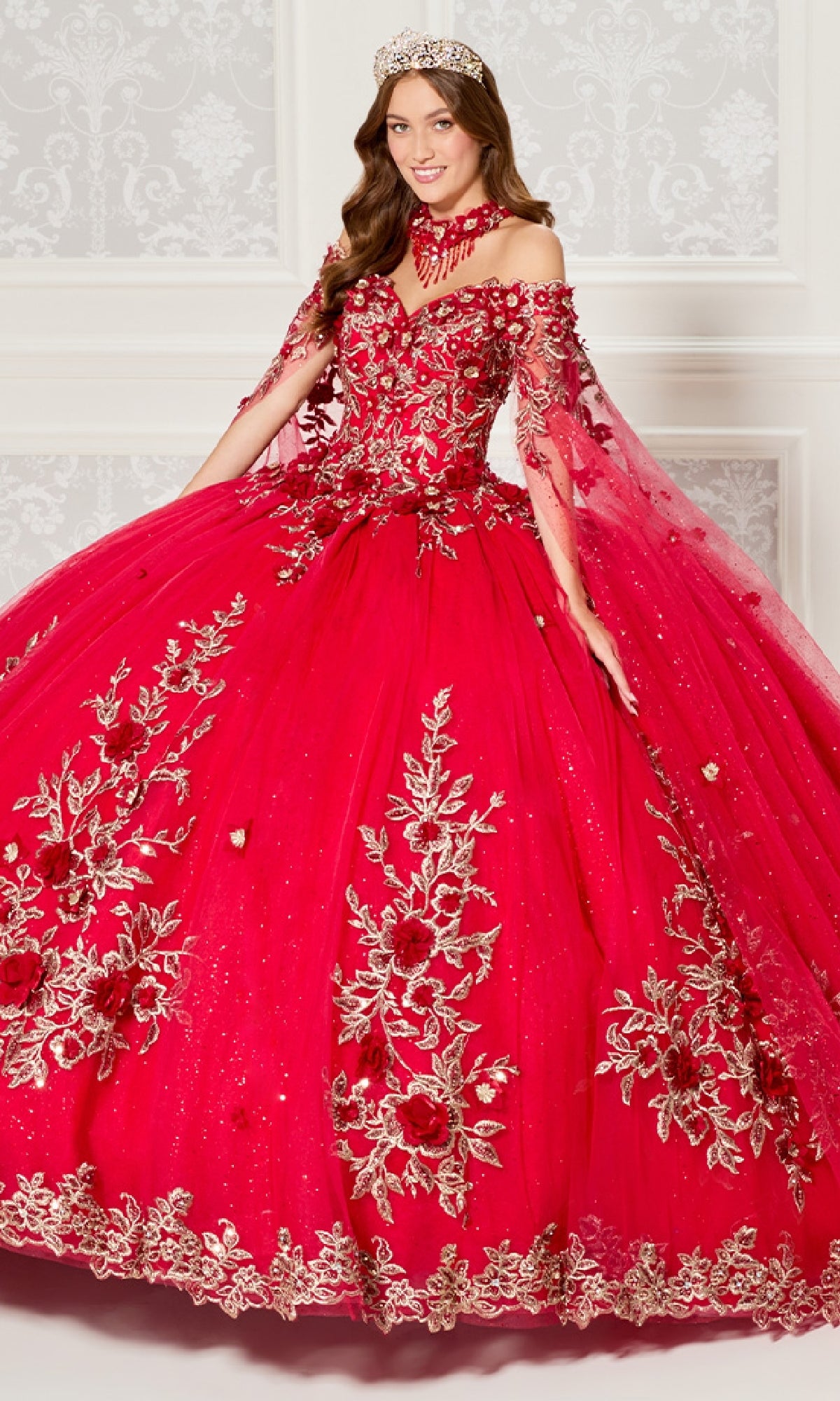 A woman stands indoors against a decorated white wall, wearing the Quinceanera Dress PR30114 By Princesa—an elaborate, bright red off-the-shoulder gown with gold floral embroidery and a matching crown.