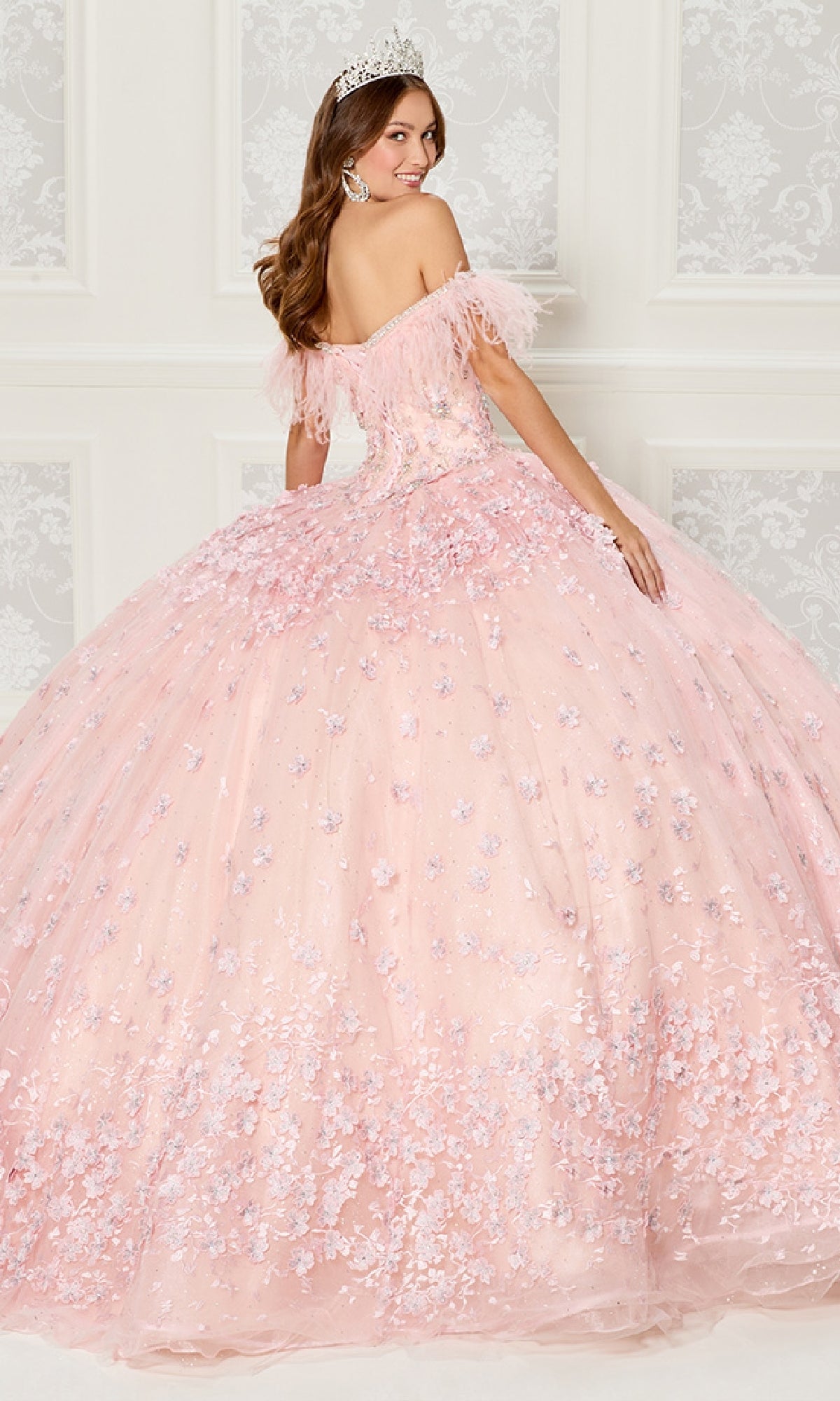 A woman in the Quinceanera Dress PR30115 By Princesa, featuring pink off-the-shoulder floral details and a tiara, stands before an ornate white wall, gazing over her shoulder.