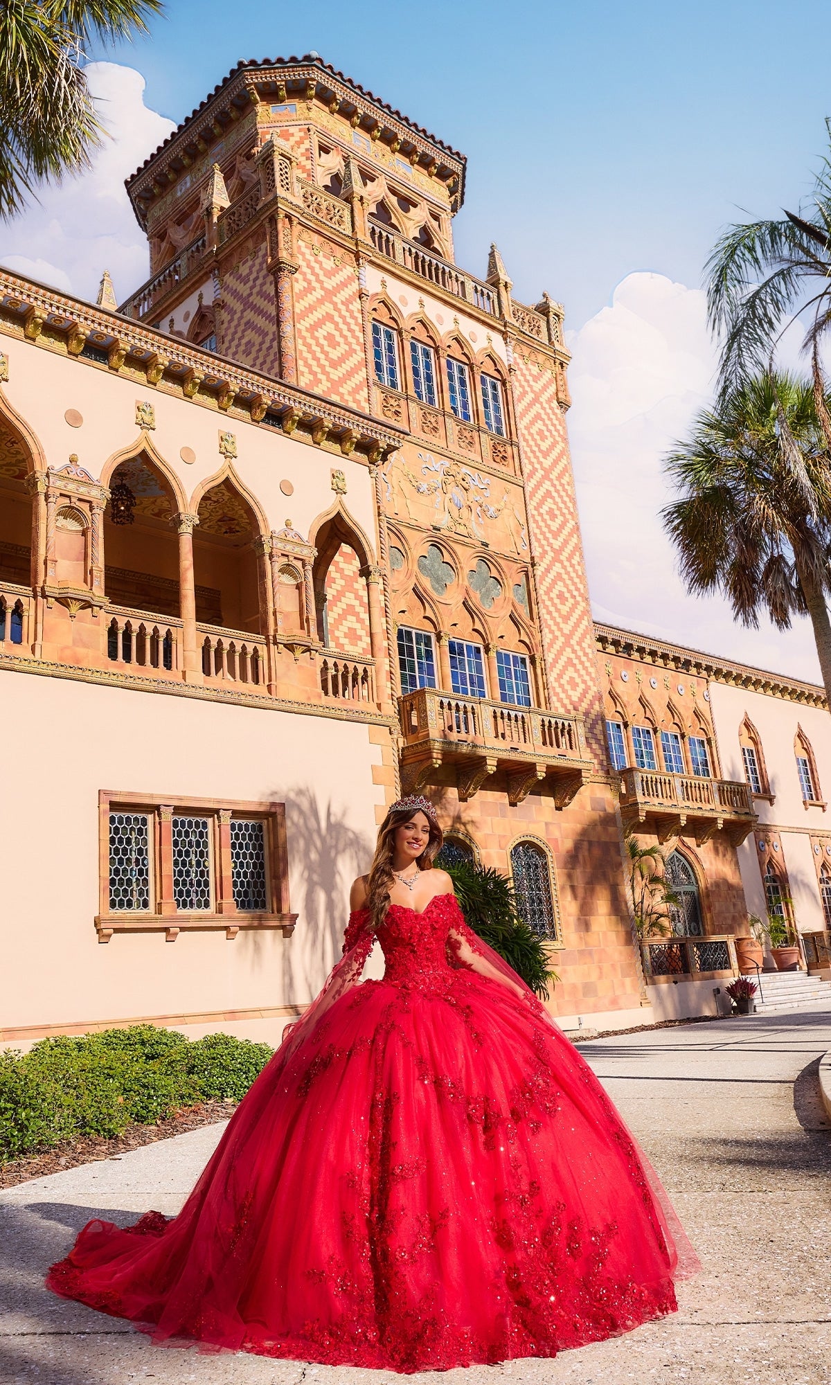 A woman wears the Quinceanera Dress PR30116 by Princesa, a stunning red ball gown with a corset bodice, standing before an ornate building with arched windows and intricate details.