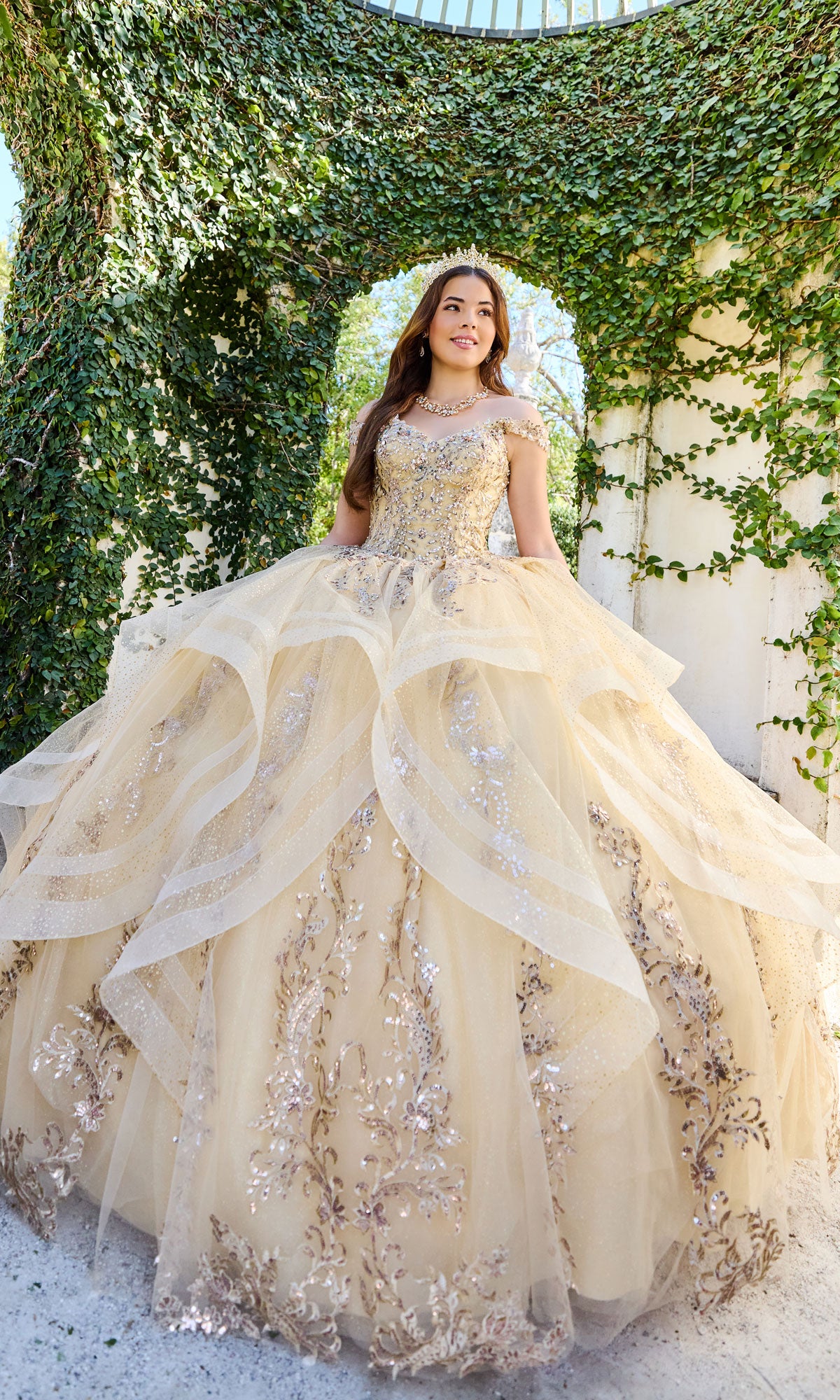 A young woman in the Quinceanera Dress PR30117 By Princesa with a detachable cape stands outdoors under an ivy-covered arch, looking to the side and smiling.
