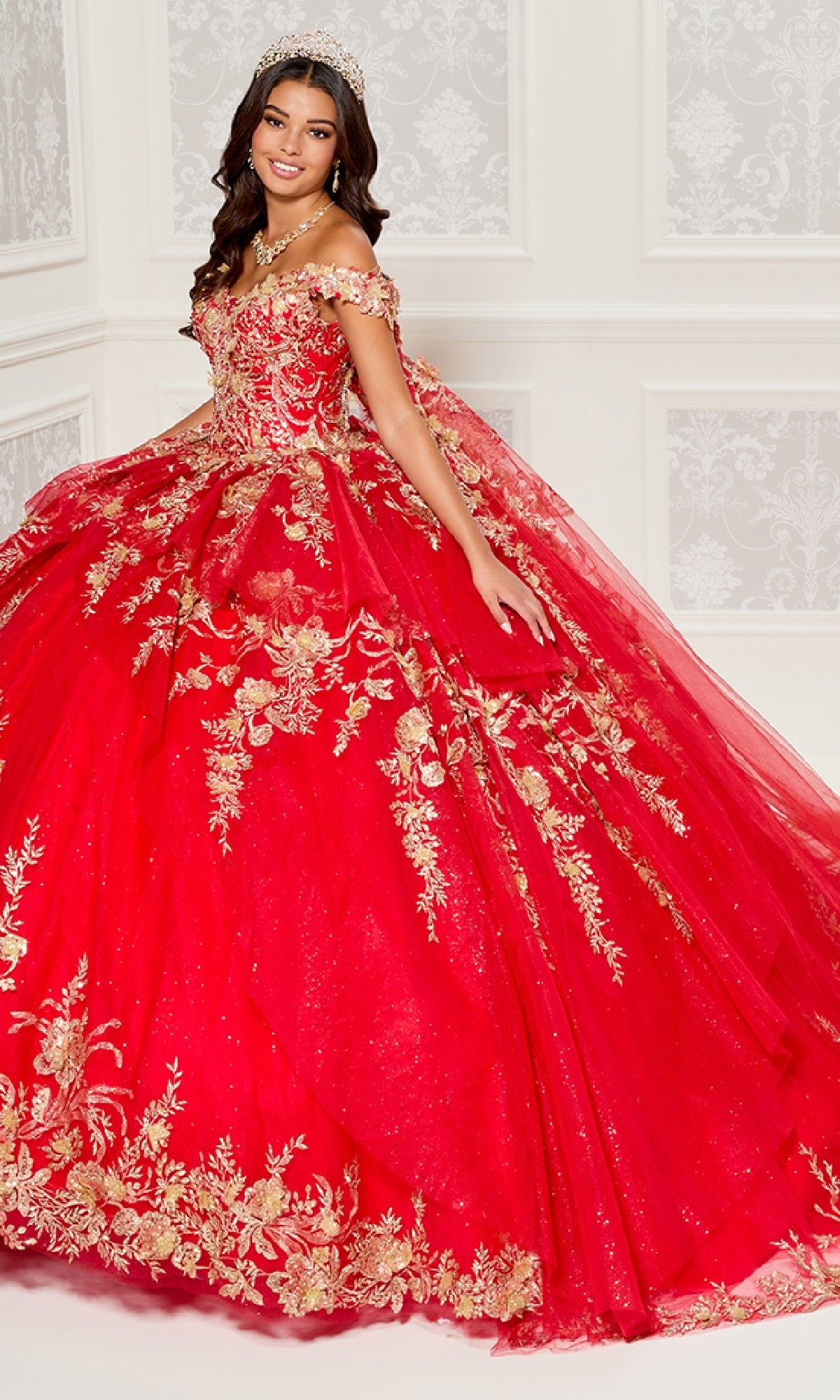 A young woman in the Quinceanera Dress PR30119 By Princesa, featuring red fabric and gold embroidery with an off-the-shoulder design and tiara, stands in an ornate white-walled room.