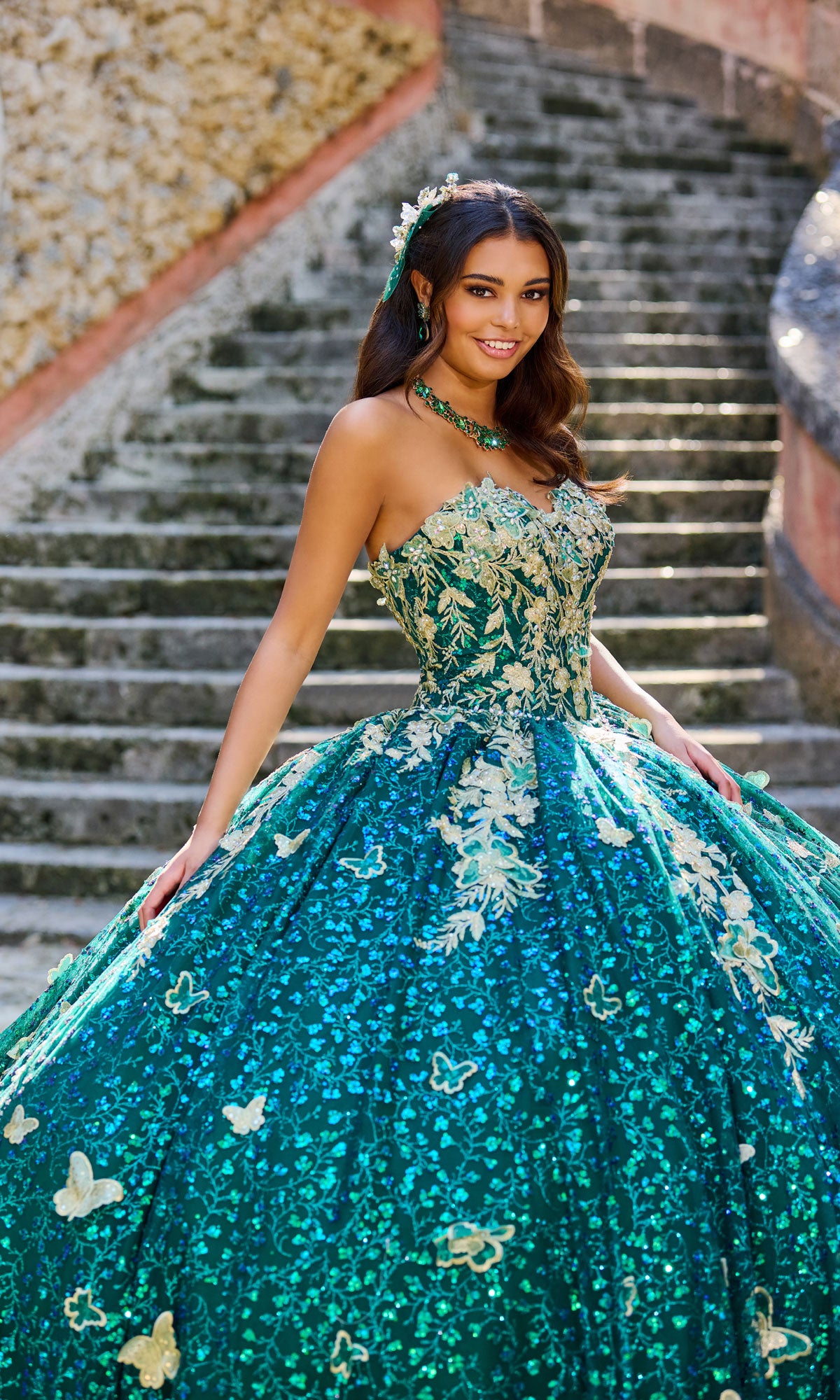 A young woman wears the Strapless Quinceanera Dress: Princesa PR30121, featuring gold floral and butterfly embroidered lace, as she stands on stone steps and smiles at the camera.